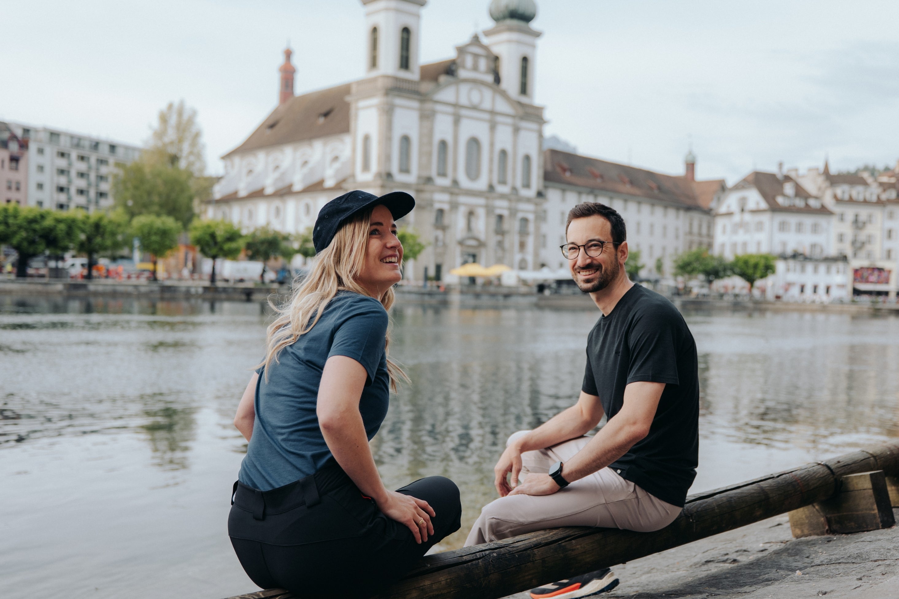 Mann und Frau in Belowzero Kleidung sitzen am Flussufer in Luzern, im Hintergrund die Jesuitenkirche