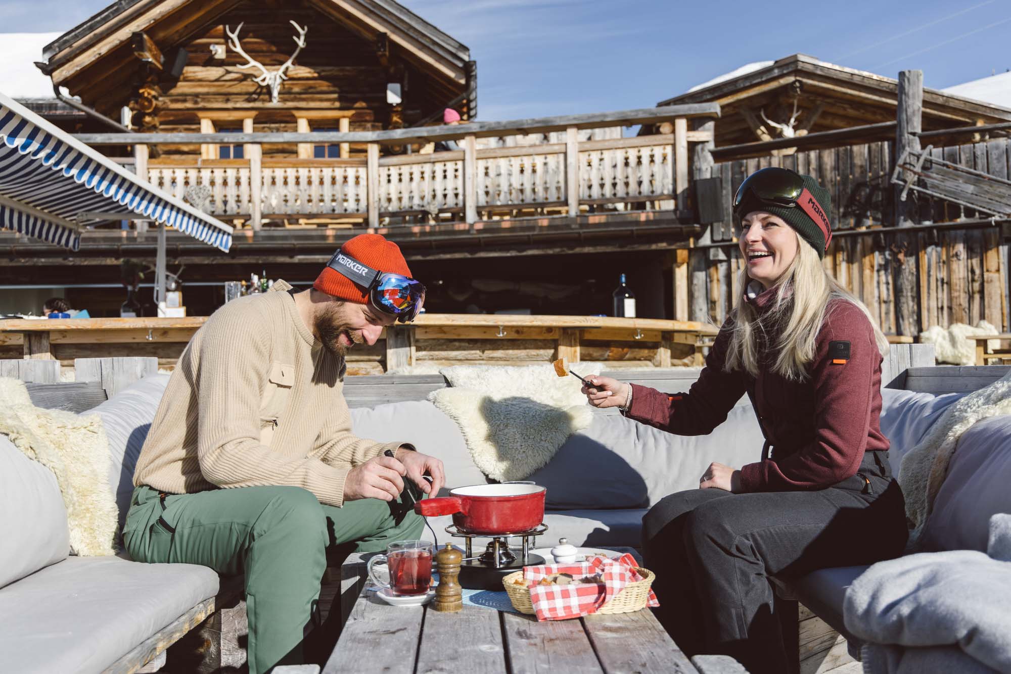 Eine Dame und ein Herr in Belowzero Bekleidung sitzen in einem Bergrestaurant und essen ein Fondue 