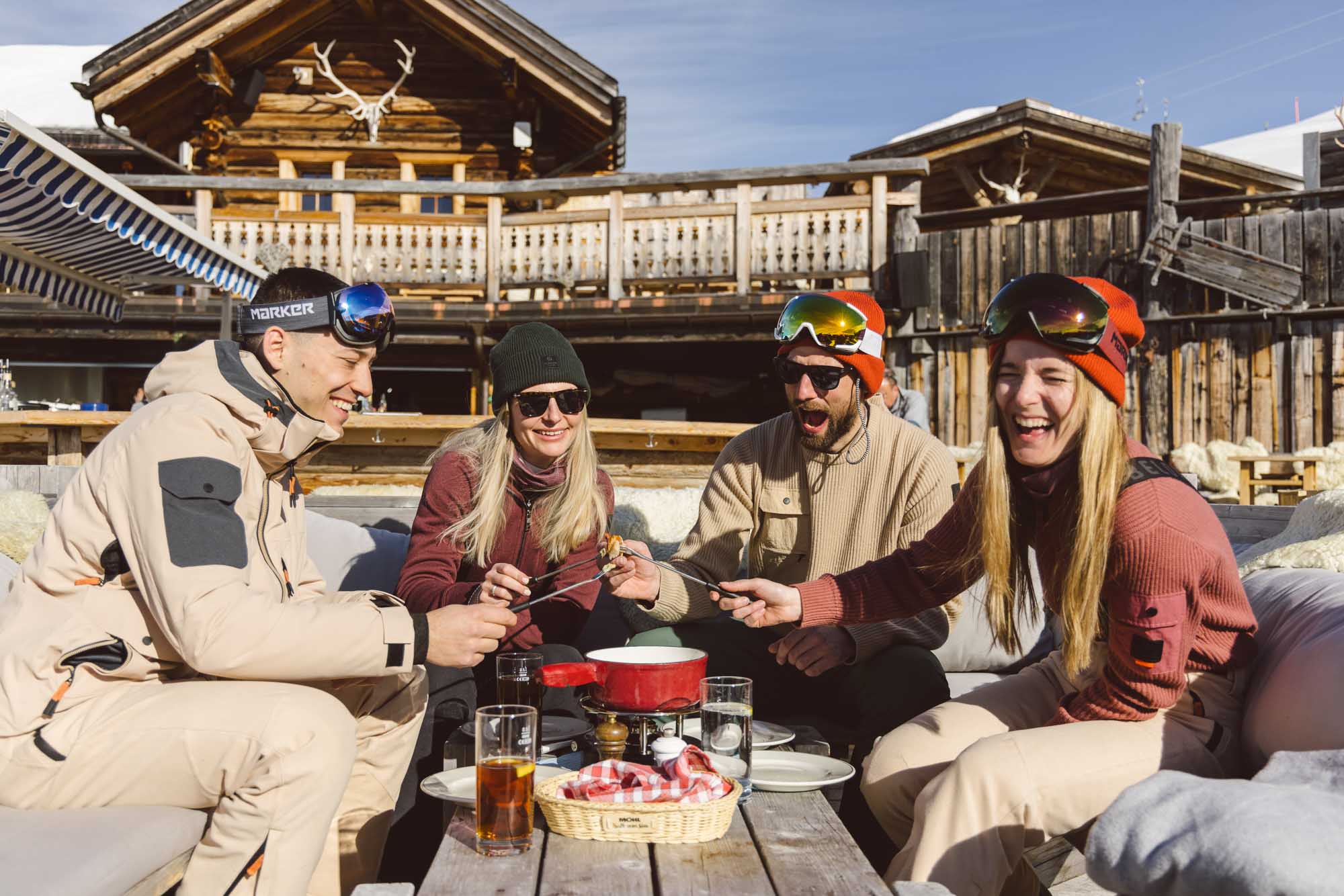 Ein Gruppe Menschen sitzt in einer Berghütte und geniesst ein Fondue. Sie tragen alle Belowzero Bekleidung.