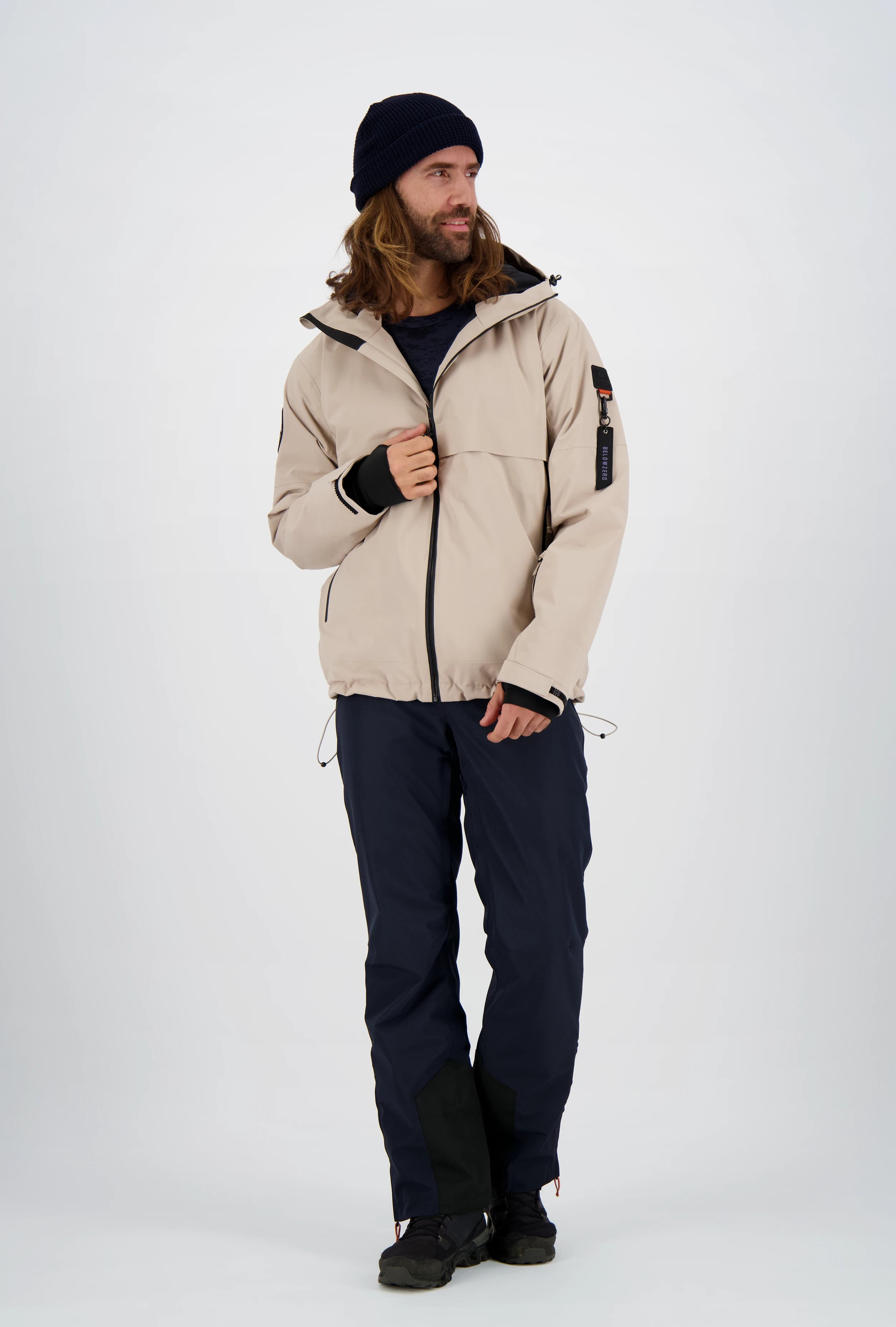 Belowzero Wintersport-Outfit für Herren: Beigefarbene Skijacke kombiniert mit dunkelblauer Funktions-Skihose und Strickmütze in voller Körperansicht.