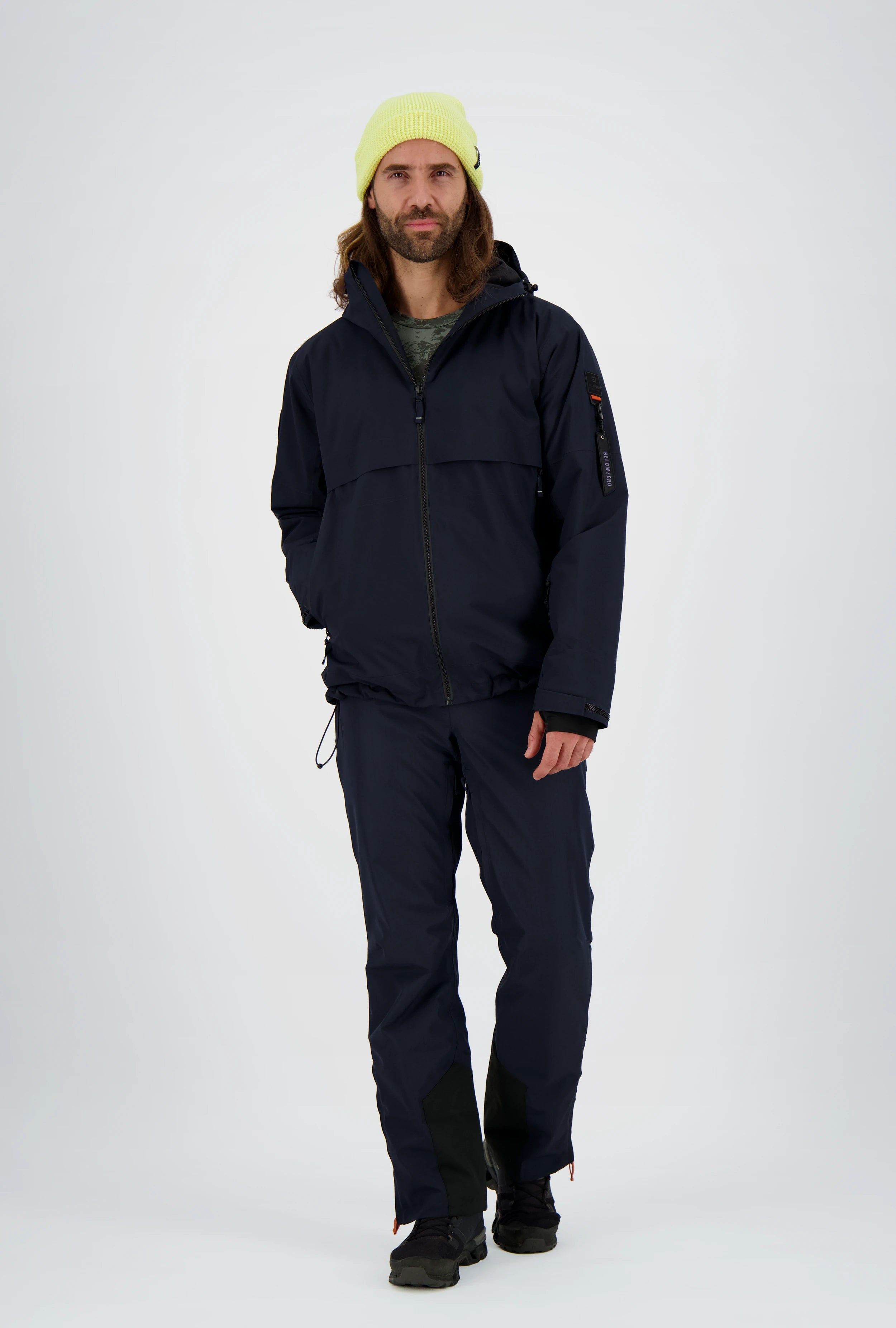 Funktionale Herren-Outdoorbekleidung von Belowzero in Dunkelblau, bestehend aus wetterfester Skijacke mit Kapuze und Ärmeltasche sowie passender Skihose mit Kantenschutz, kombiniert mit neongelber Strickmütze und schwarzen Winterschuhen.