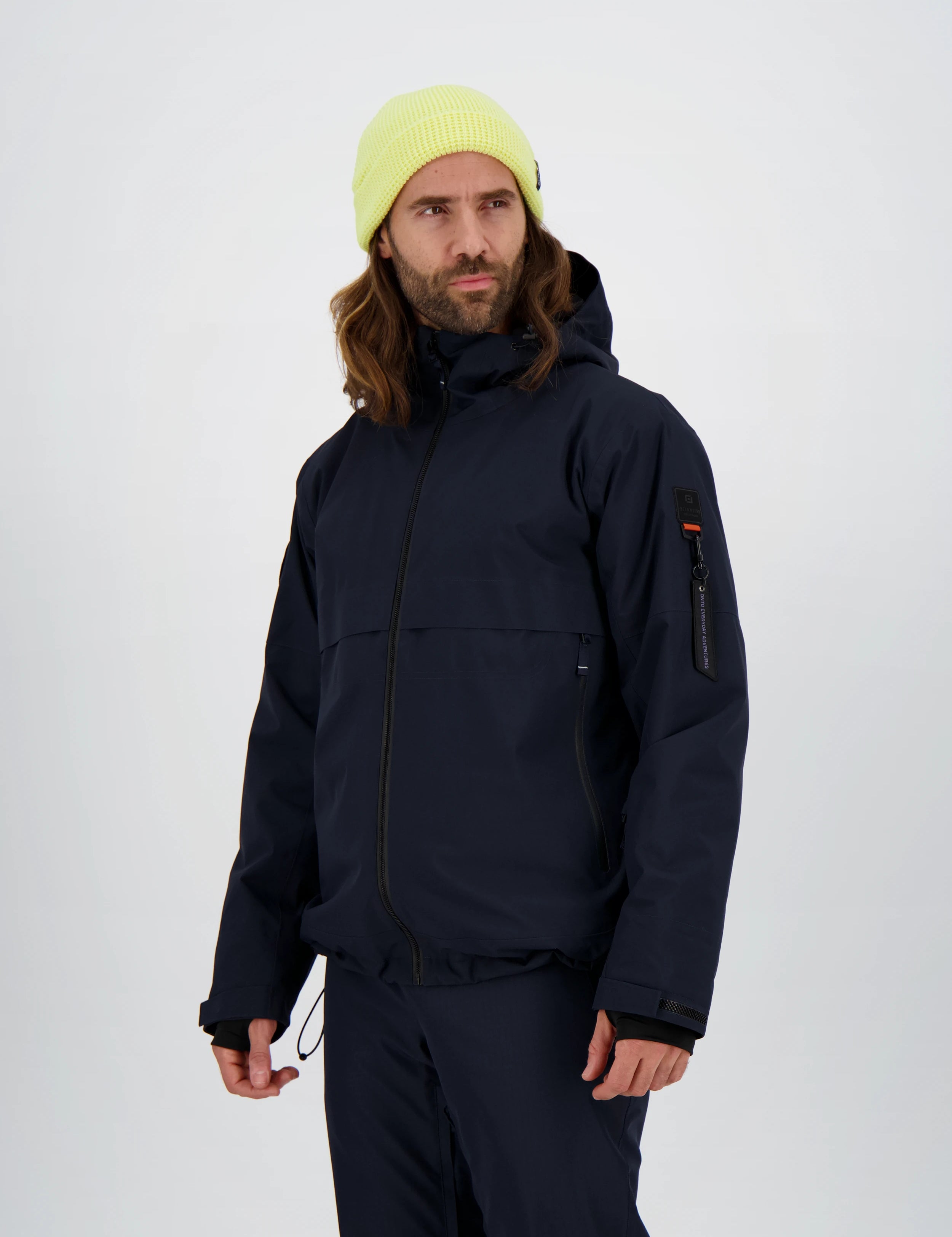 Dunkelblaue Herren-Skijacke von Belowzero mit Kapuze, seitlichen Reissverschlusstaschen und markanter Ärmeltasche inklusive Zippuller, kombiniert mit passender Skihose und neongelber Strickmütze.