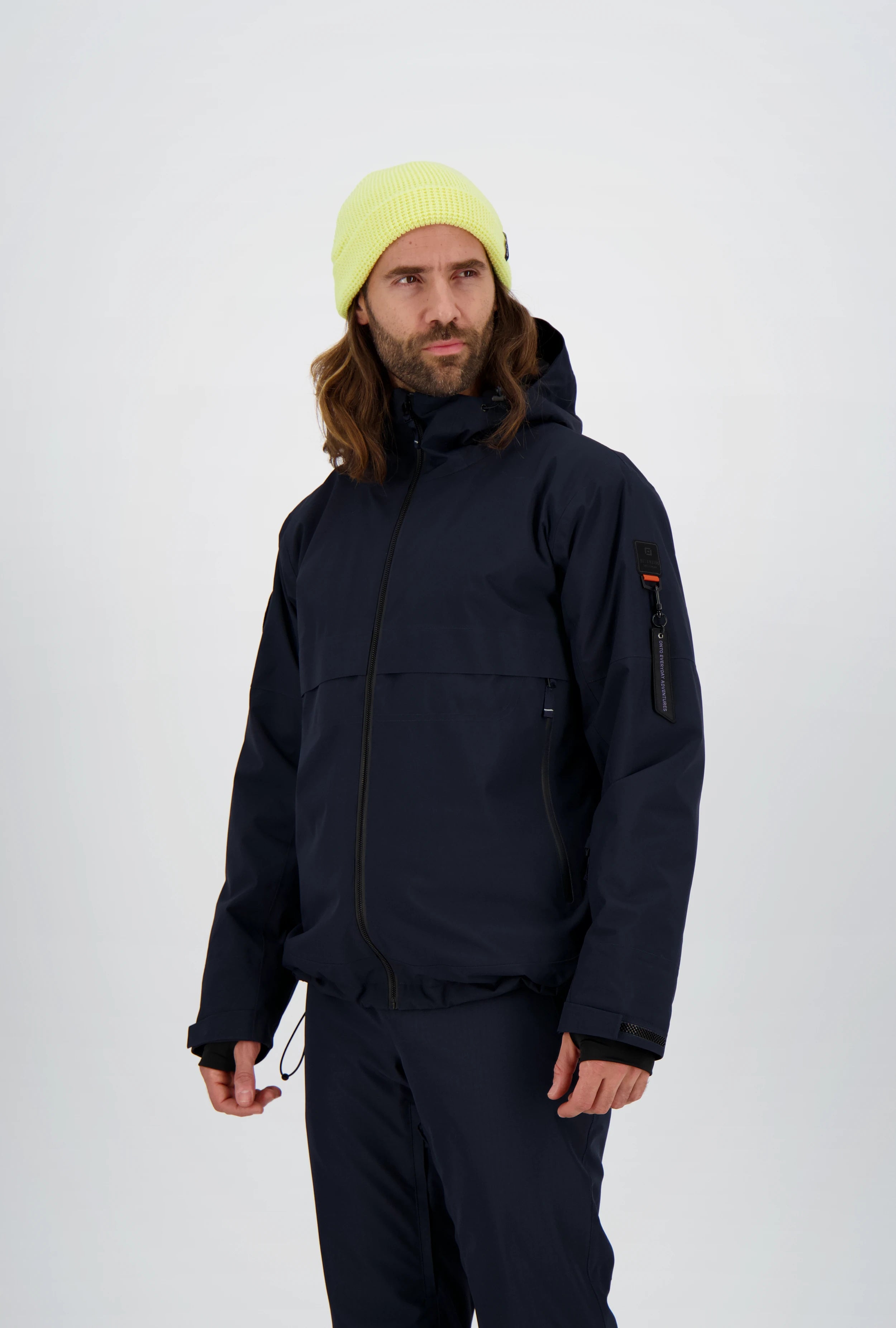 Dunkelblaue Herren-Skijacke von Belowzero mit Kapuze, seitlichen Reissverschlusstaschen und markanter Ärmeltasche inklusive Zippuller, kombiniert mit passender Skihose und neongelber Strickmütze.