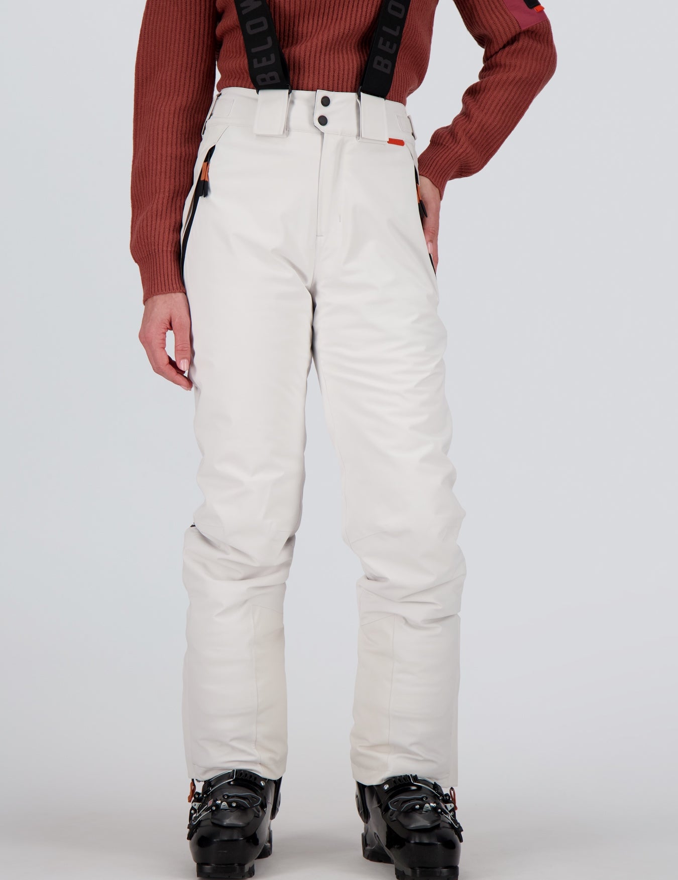 Frontansicht einer weissen Damen Skihose Andermatt von Belowzero Weisse Damen-Skihose der Schweizer Marke Belowzero mit verstellbaren schwarzen Hosenträgern samt Logo-Schriftzug. Die Hose bietet seitliche Reißverschlusstaschen mit orangefarbenen Akzenten und wird zusammen mit schwarzen Skischuhen sowie einem rostbraunen Strickpullover präsentiert