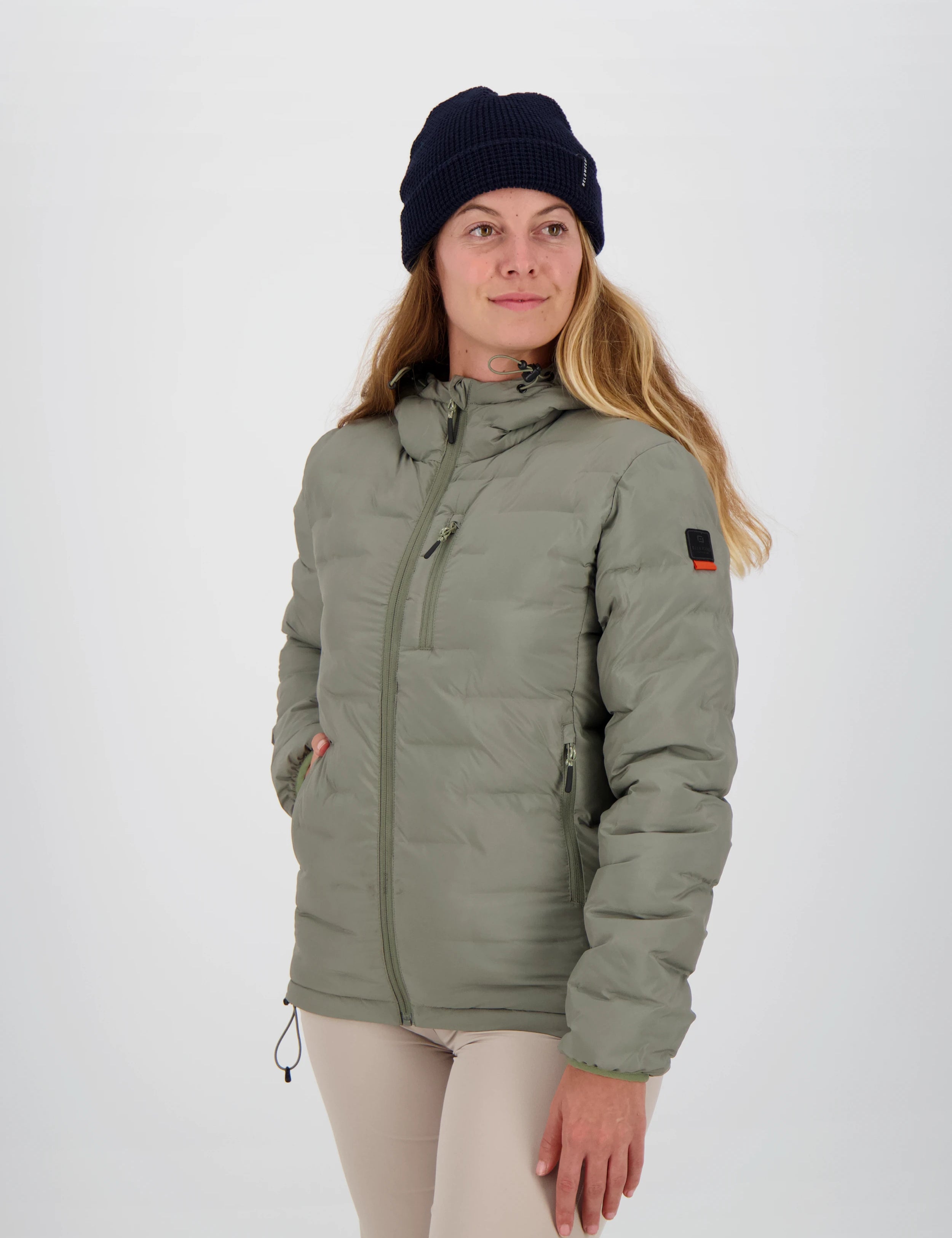 Gesteppte salbeigrüne grüne Damen-Outdoorjacke von Belowzero mit Kapuze, Reissverschlusstaschen und Logo-Badge am Ärmel, getragen mit beiger Funktionshose und dunkelblauer Strickmütze.