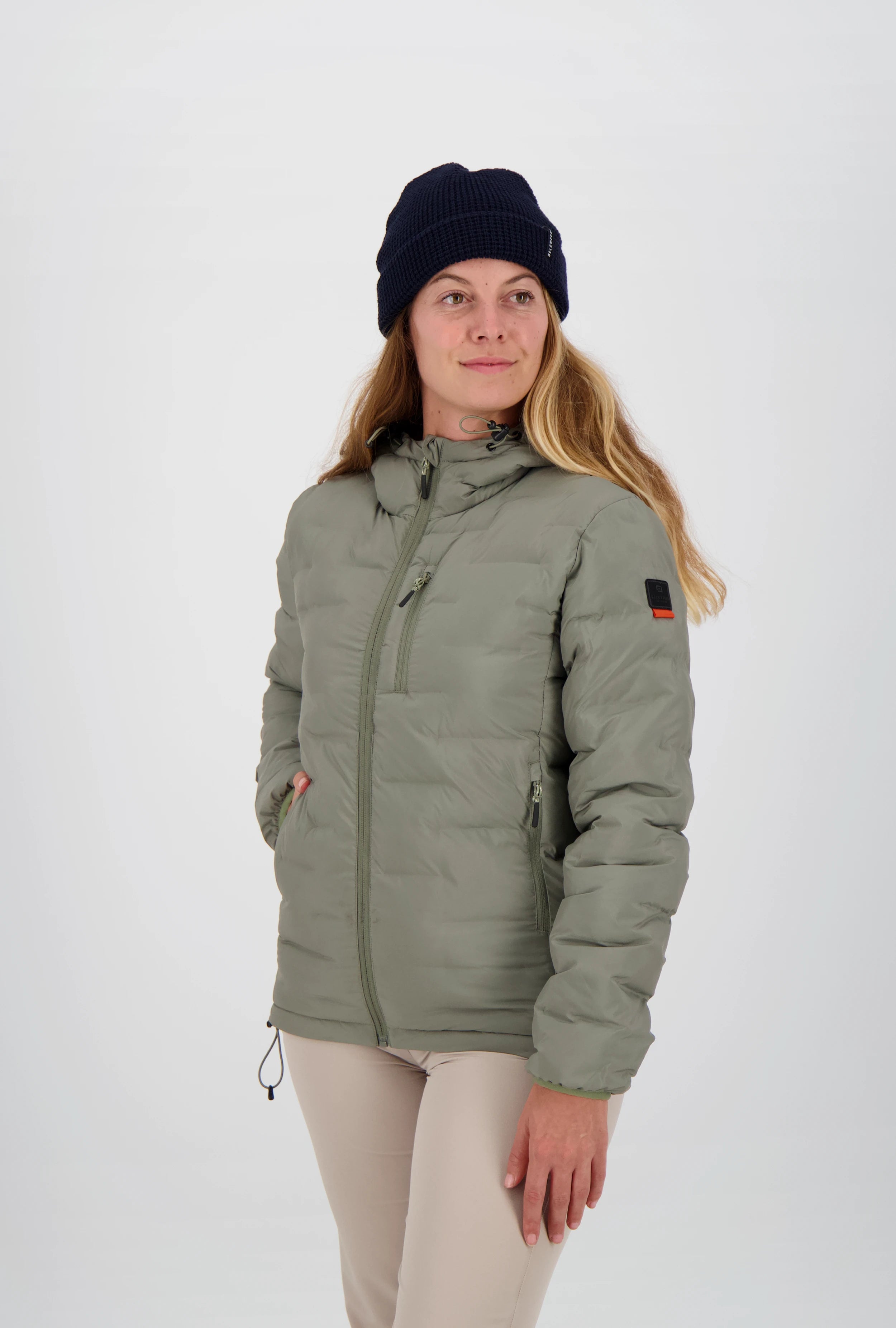 Gesteppte salbeigrüne grüne Damen-Outdoorjacke von Belowzero mit Kapuze, Reissverschlusstaschen und Logo-Badge am Ärmel, getragen mit beiger Funktionshose und dunkelblauer Strickmütze.
