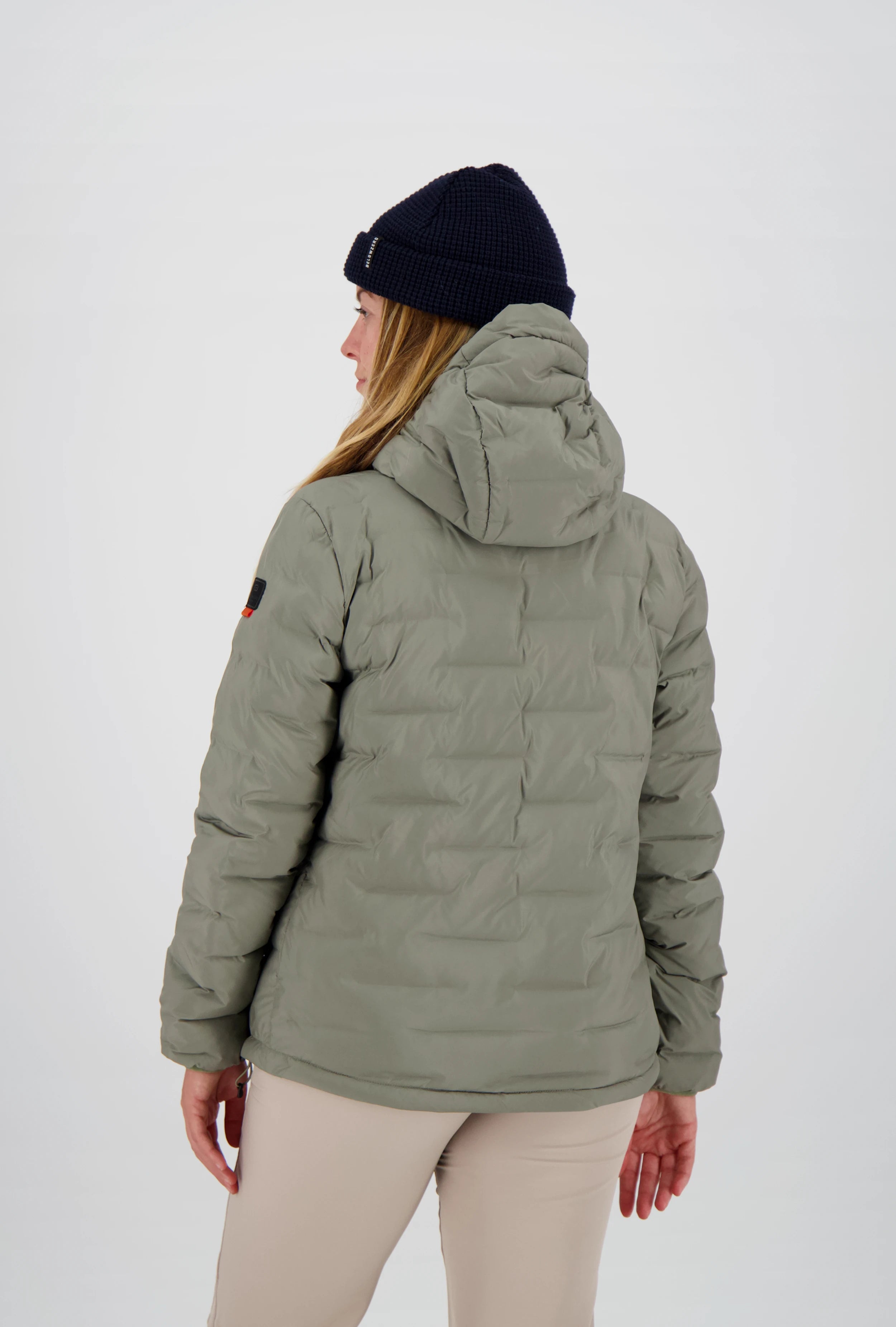 Rückansicht einer gesteppten salbeigrünen Damen-Outdoorjacke von Belowzero mit verstellbarer Kapuze und Logo-Badge am linken Ärmel, getragen mit beiger Funktionshose und dunkelblauer Beanie.