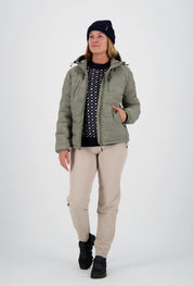 Geöffnete salbeigrüne Damen-Steppjacke von Belowzero mit Kapuze über einem dunkelblauen Muster-Swissness Strickpullover, getragen mit beiger Funktionshose, schwarzer Beanie und schwarzen Outdoorschuhen.
