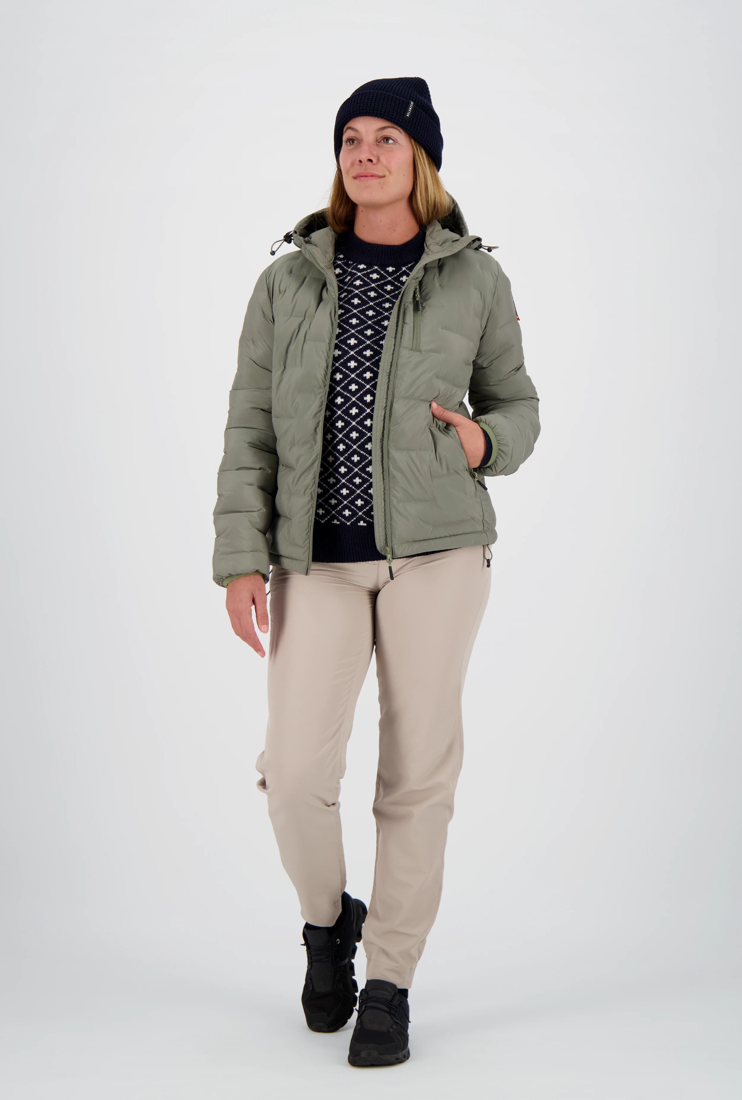 Geöffnete salbeigrüne Damen-Steppjacke von Belowzero mit Kapuze über einem dunkelblauen Muster-Swissness Strickpullover, getragen mit beiger Funktionshose, schwarzer Beanie und schwarzen Outdoorschuhen.