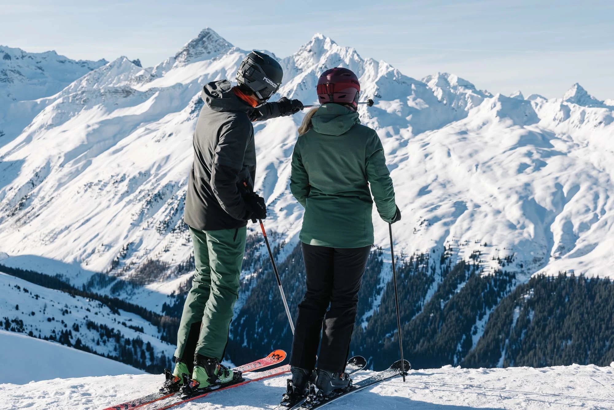 Eine Dame und ein Herr gekleidet in Belowzero schauen von einer Piste auf die Winterberglandschaft