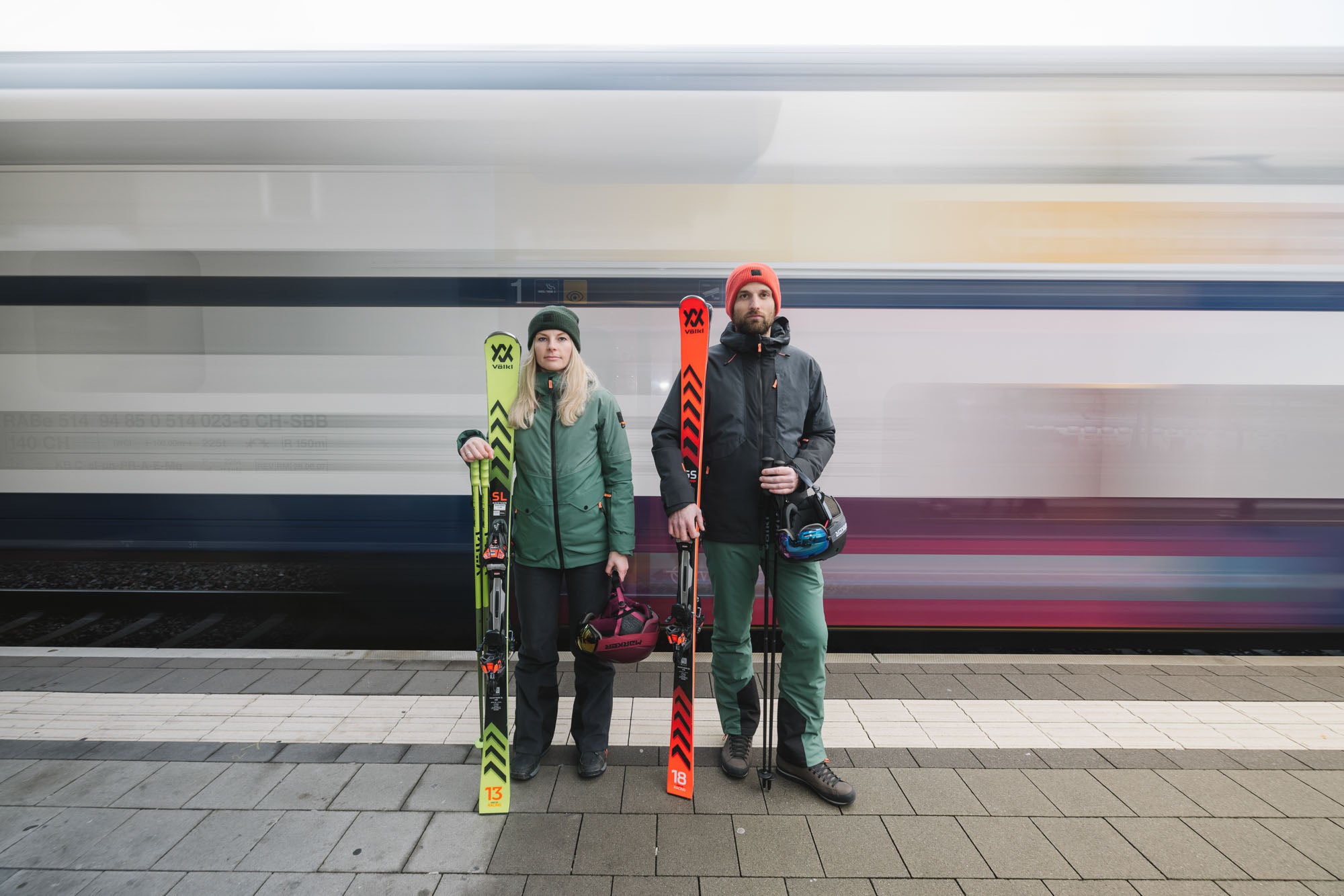 EIne Dame und ein Herr in Belowzero Bekleidung stehen mit Skiausrüstung am Bahnhof