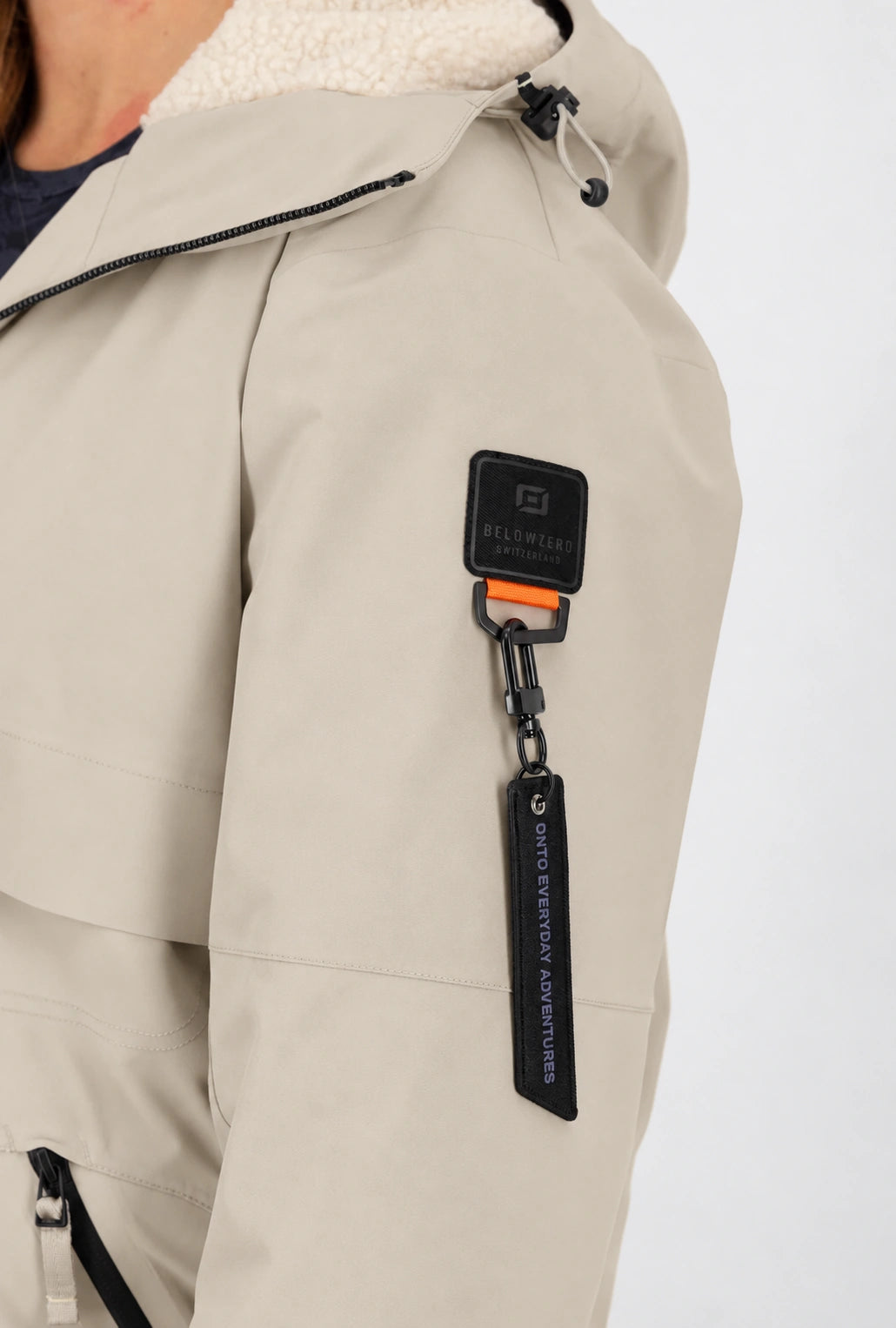 Detailansicht der sandfarbenen Belowzero Damen-Outdoorjacke mit Teddyfell-Kapuze, Oberarm-Badge und «Onto Everyday Adventures»-Karabineranhänger.