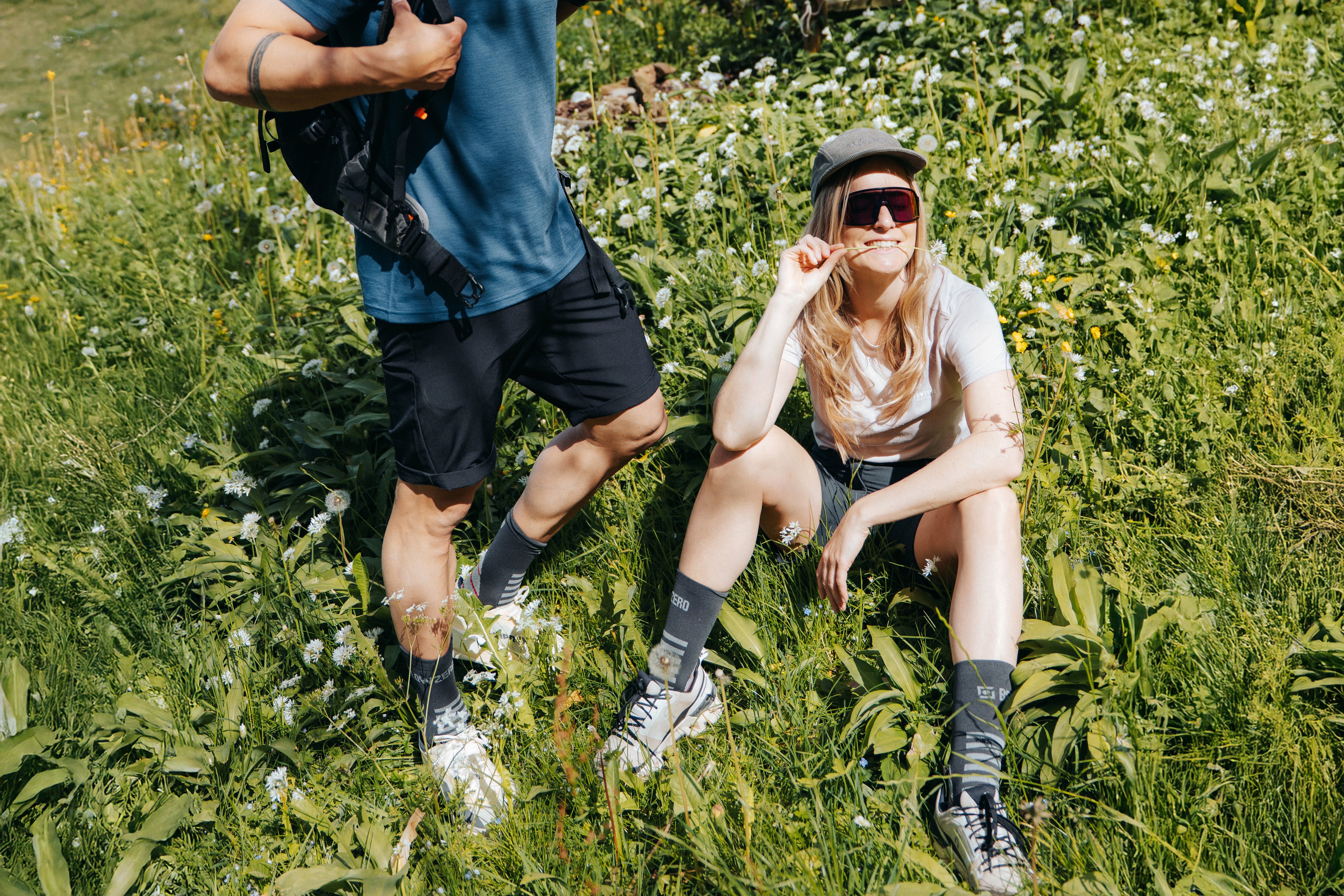 Ein Mann und eine Frau in funktionaler Belowzero Outdoor-Sommerkleidung sitzen auf Sommerwiese und tragen Belowzero Outdoorkleidung Zu sehen sind ein blaues T-Shirt, schwarze Wandershorts, ein hellrosa Damen-T-Shirt, graue Outdoor-Socken Trekkingsocken und eine Kappe.
