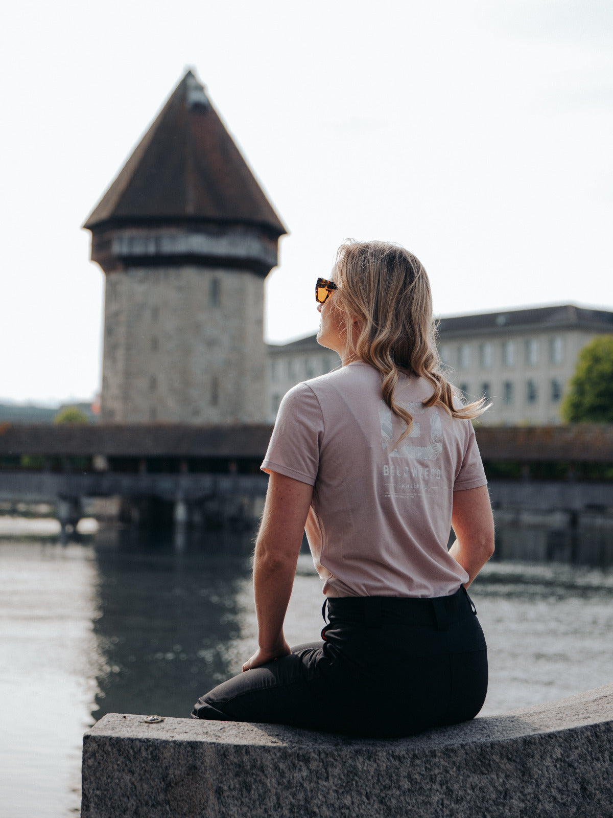Frau in Belowzero rosa Funktionsshirt sitzt am Ufer der Reuss in Luzern, im Hintergrund der Wasserturm. ro