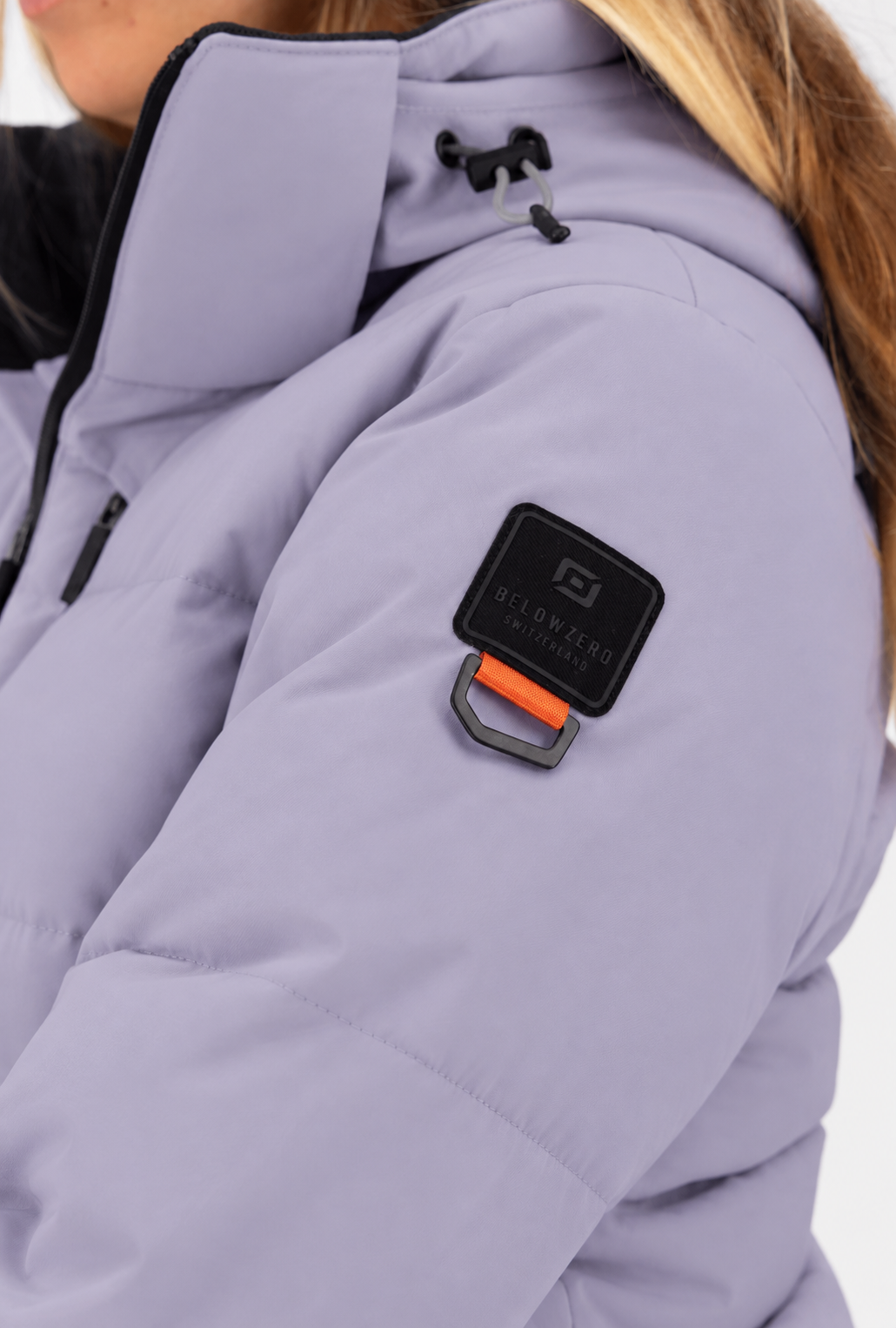 Belowzero Skijacke für Damen in Flieder violett lila – Detailansicht des Oberarm-Logos mit Belowzero Switzerland Badge, D-Ring und verstellbarer Kapuze.