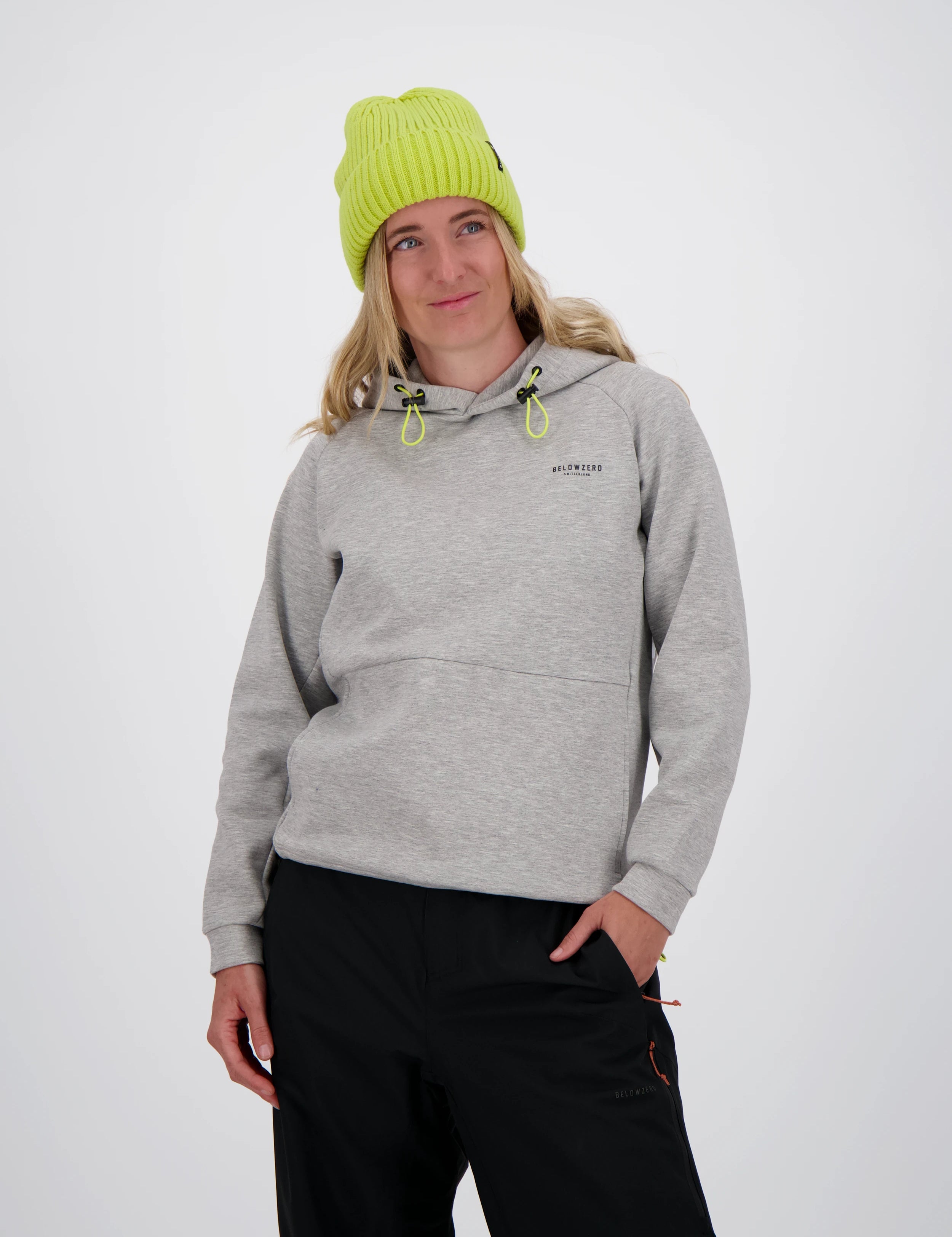 Frau mit neongelber Beanie-Mütze trägt einen grauen Belowzero Hoodie mit Kapuze und gelben Kordelzügen sowie eine schwarze Outdoorhose.