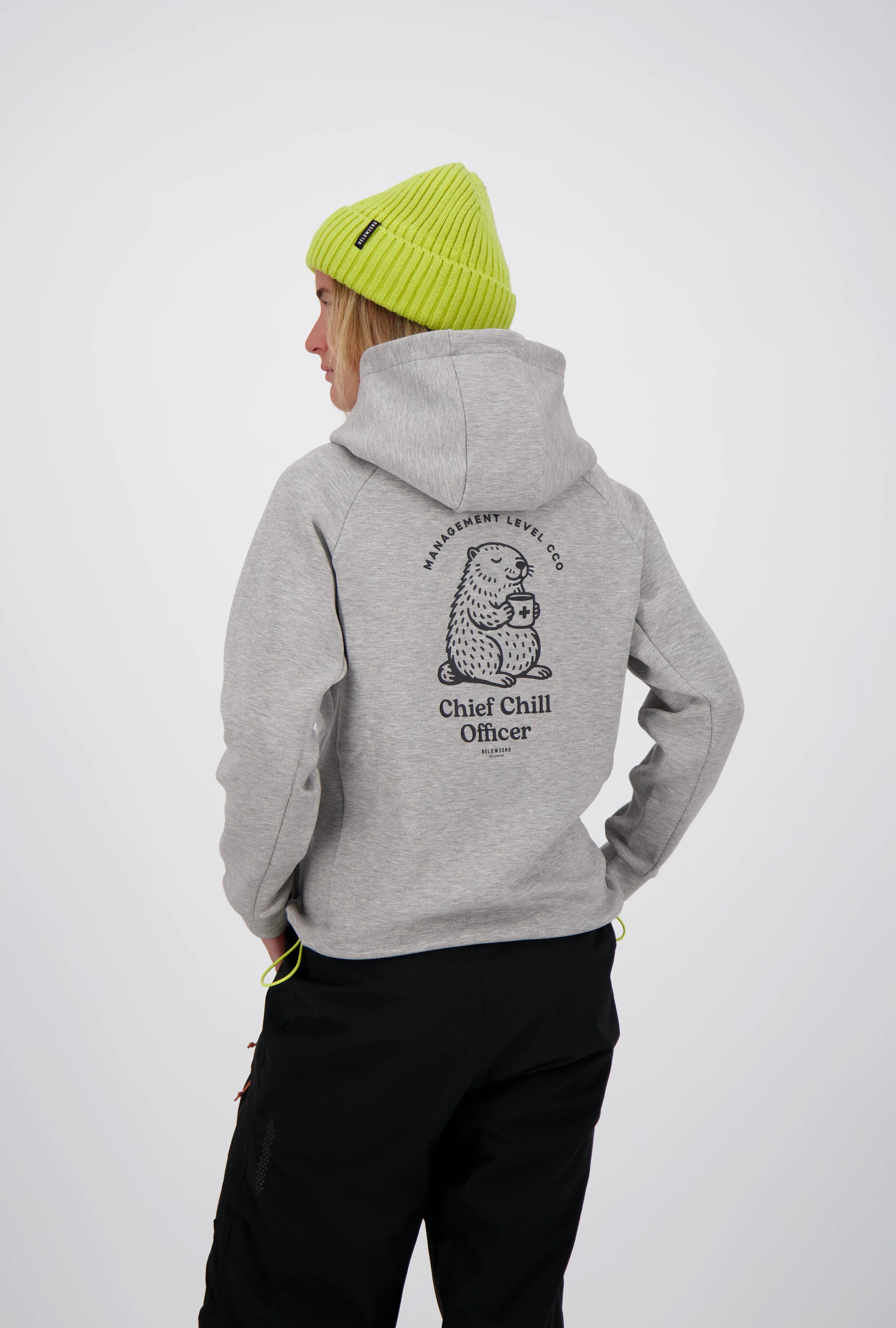 Rückenansicht einer Frau im grauen Belowzero Kapuzenpullover mit Murmeltier-Print "Chief Chill Officer", neongelber Beanie-Mütze und schwarzer Outdoorhose.