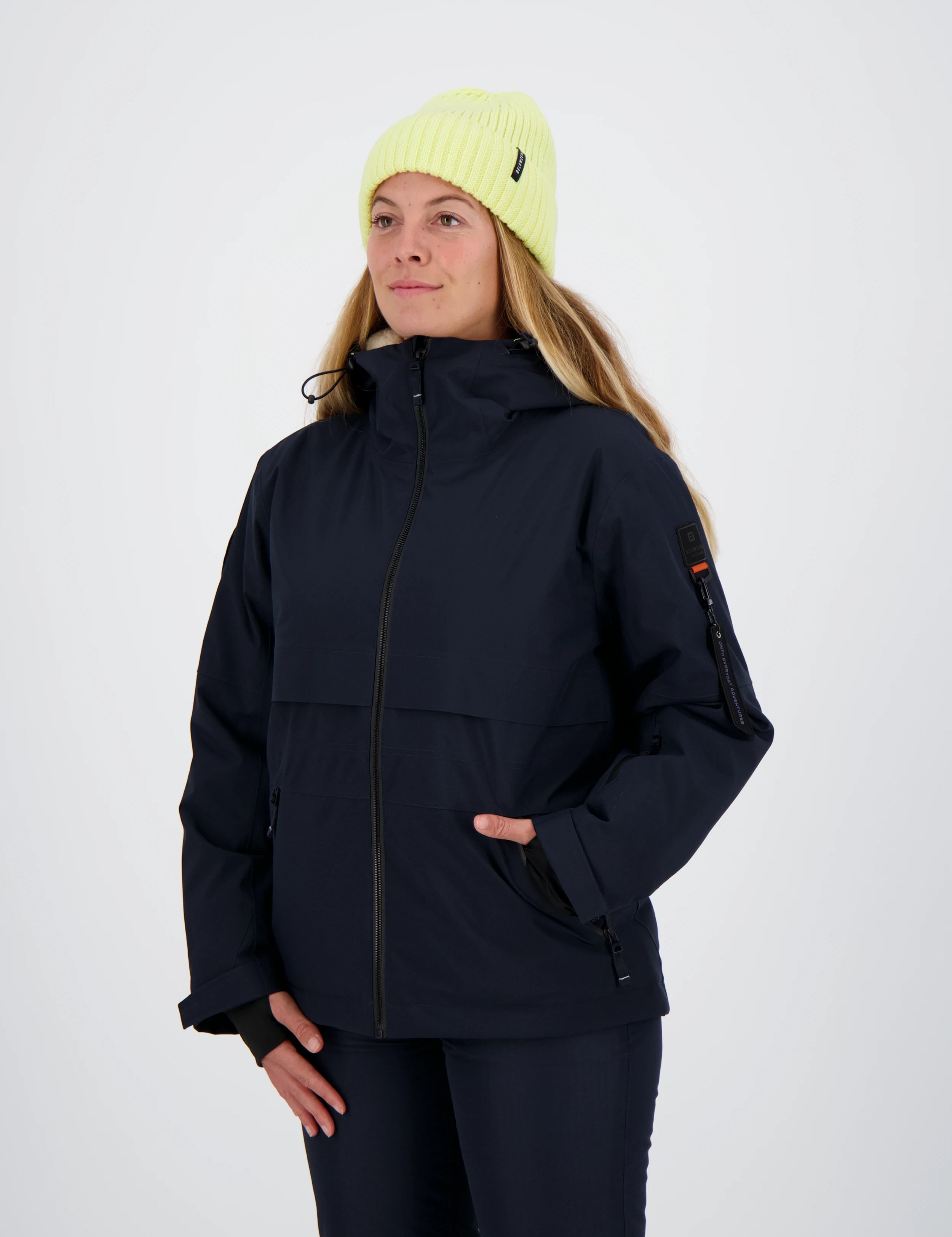 Frau mit neongelber Beanie-Mütze trägt eine dunkelblaue Belowzero Damen Outdoorjacke mit Kapuze, Ärmeltasche Pulswärmer und passender dunkler Hose.