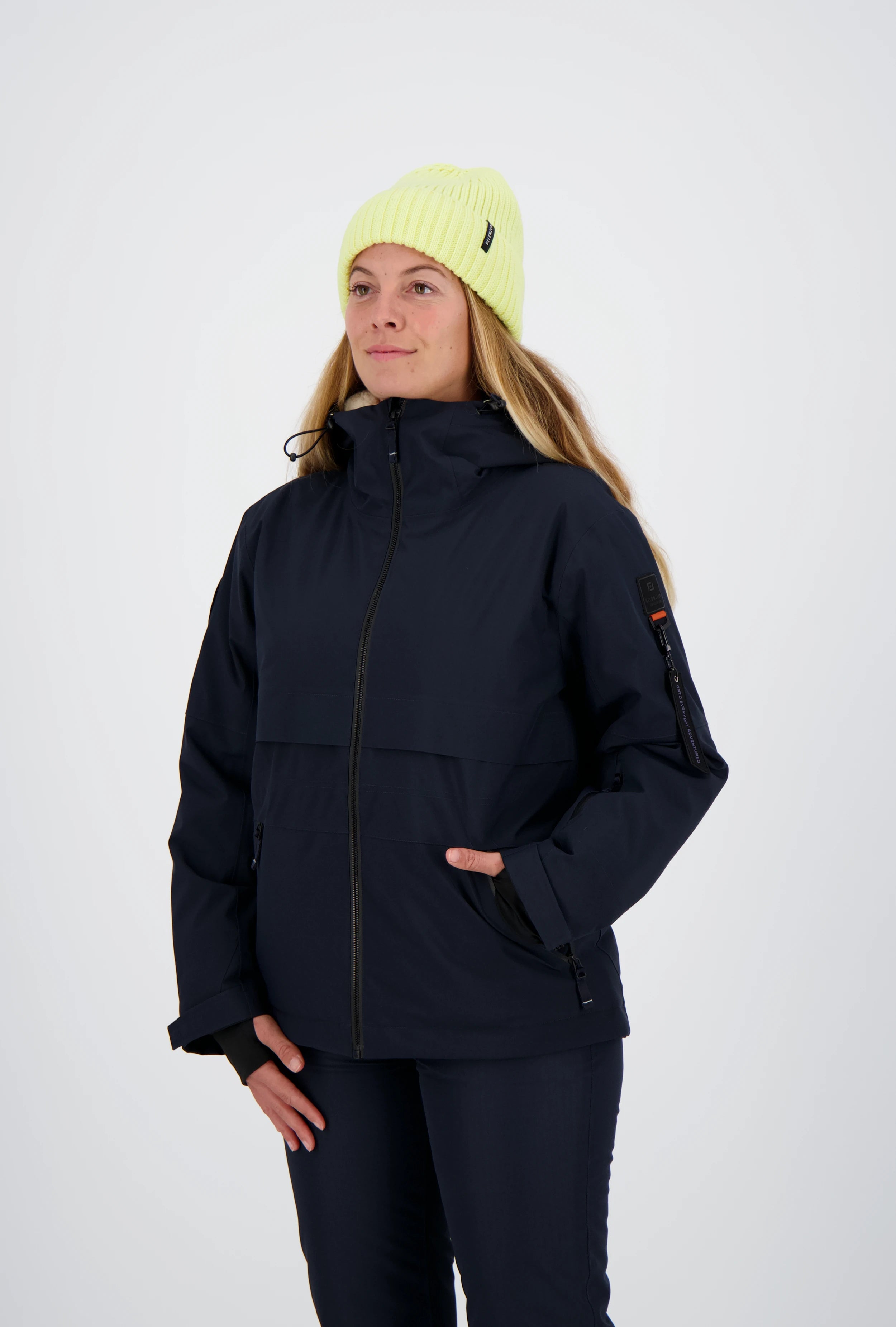 Frau mit neongelber Beanie-Mütze trägt eine dunkelblaue Belowzero Damen Outdoorjacke mit Kapuze, Ärmeltasche Pulswärmer und passender dunkler Hose.