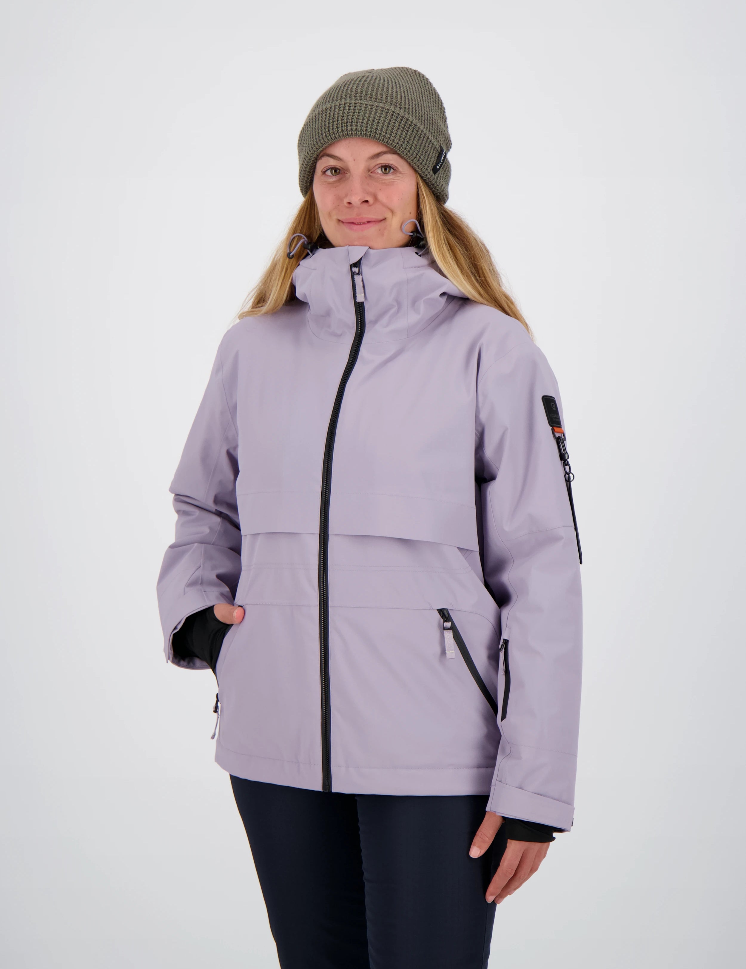 Frontansicht einer Frau mit olivgrüner Beanie-Mütze in einer fliederfarbenen Belowzero Damen Outdoorjacke mit hochgeschlossenem Kragen und Seitentaschen.