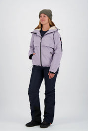 Frau mit olivgrüner Beanie-Mütze trägt eine fliederfarbene Belowzero Damen Outdoorjacke mit Fellfutter-Kapuze, dunkelblaue Skihose und schwarze Sportschuhe.
