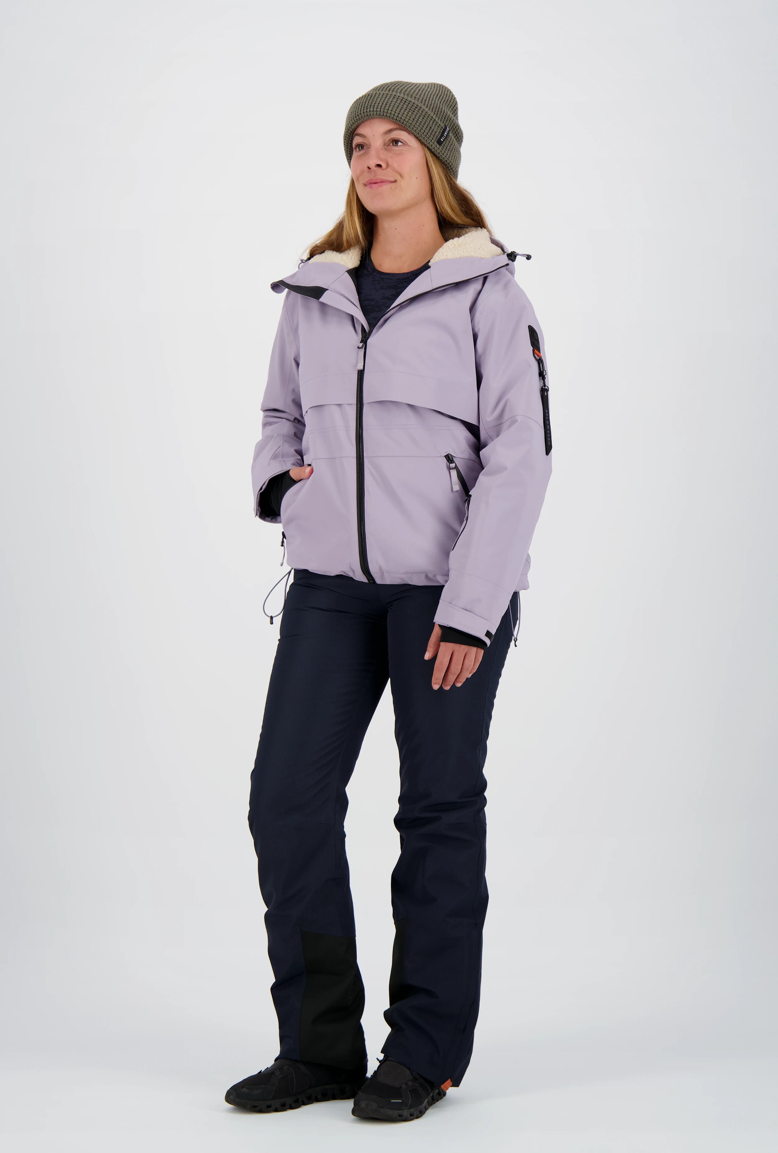 Frau mit olivgrüner Beanie-Mütze trägt eine fliederfarbene Belowzero Damen Outdoorjacke mit Fellfutter-Kapuze, dunkelblaue Skihose und schwarze Sportschuhe.
