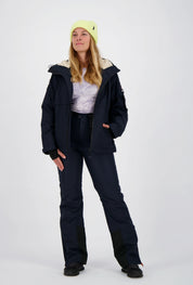 Frau mit neongelber Beanie-Mütze trägt eine geöffnete dunkelblaue Belowzero Outdoorjacke Skijacke mit helles Fellfutter in der Kapuze, passende dunkelblaue Skihose und schwarze Sportschuhe.