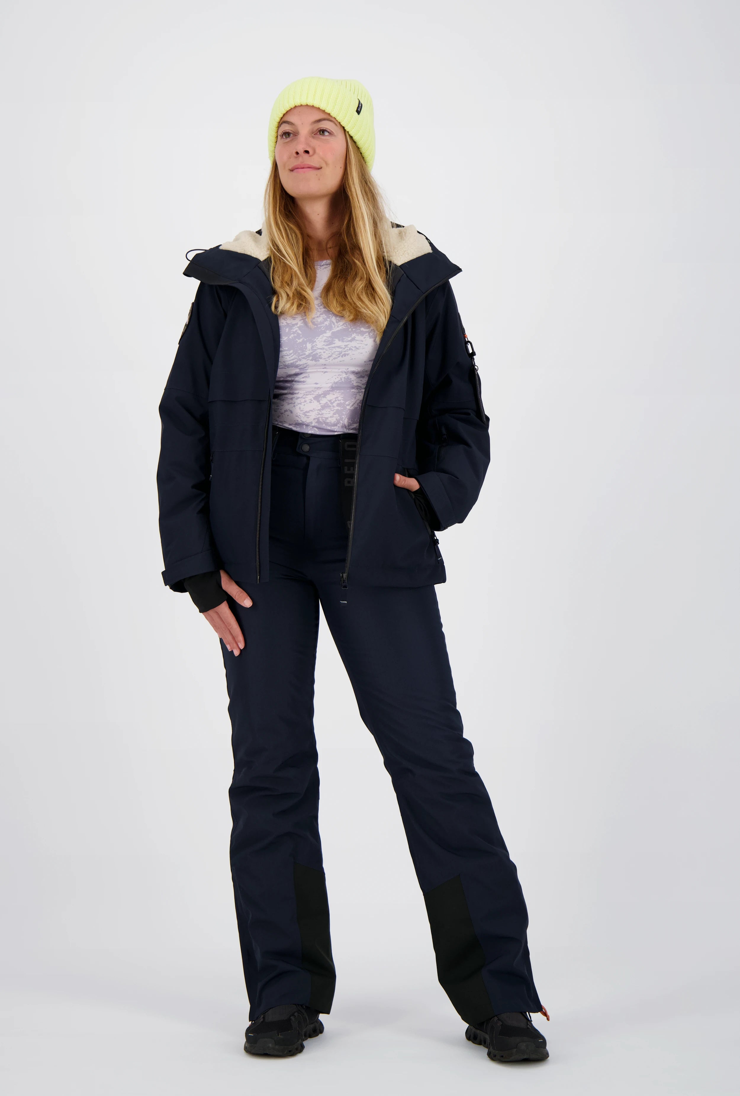 Frau mit neongelber Beanie-Mütze trägt eine geöffnete dunkelblaue Belowzero Outdoorjacke Skijacke mit helles Fellfutter in der Kapuze, passende dunkelblaue Skihose und schwarze Sportschuhe.