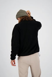 Rückenansicht eines Mannes mit langen Haaren und olivgrüner Beanie-Mütze, der eine schwarze Belowzero Teddyfleece-Jacke und eine helle Outdoorhose trägt.