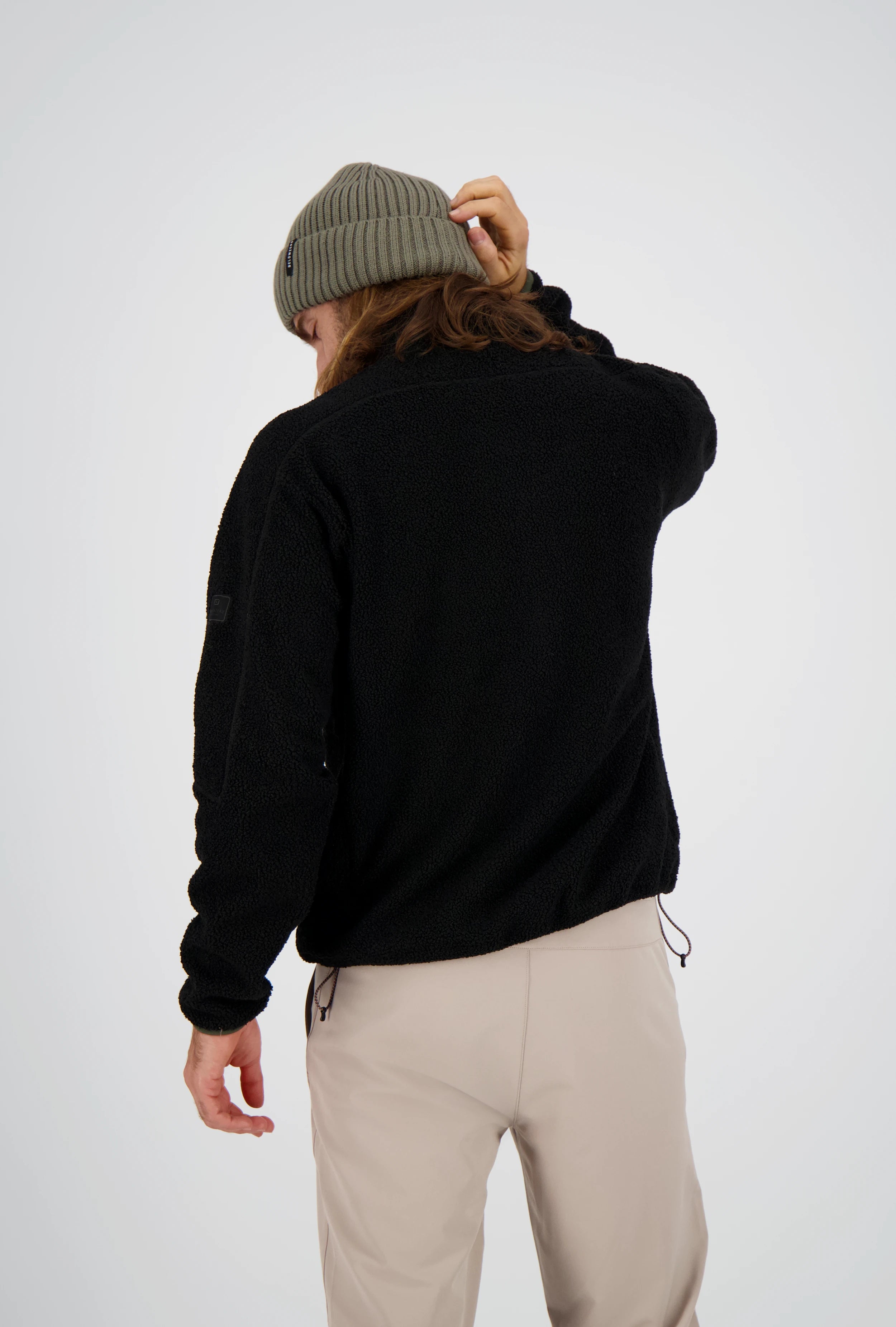 Rückenansicht eines Mannes mit langen Haaren und olivgrüner Beanie-Mütze, der eine schwarze Belowzero Teddyfleece-Jacke und eine helle Outdoorhose trägt.