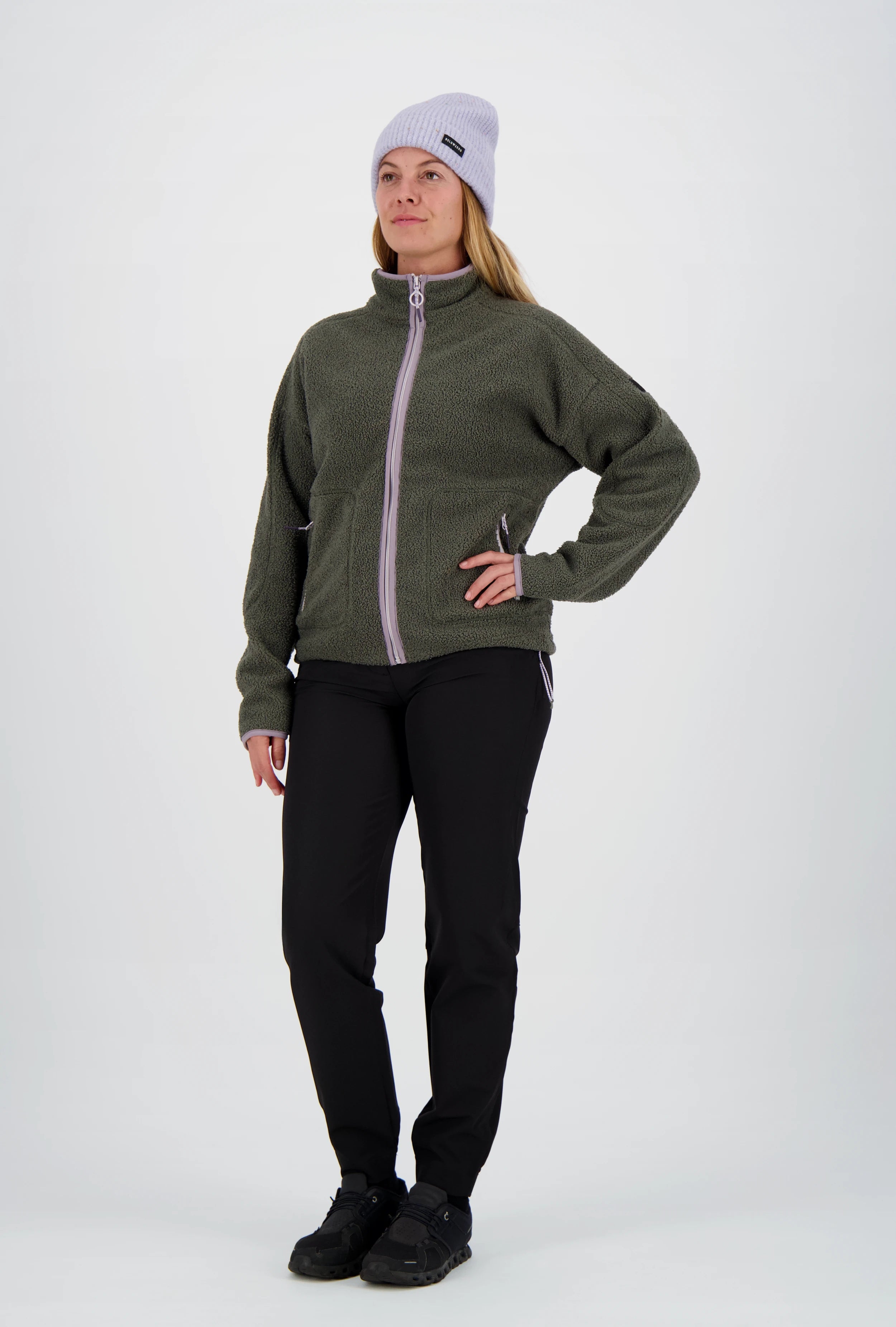 Frau mit fliederfarbener Beanie-Mütze trägt eine dunkelgrüne Belowzero Teddyfleece-Jacke mit fliederfarbenem Reissverschluss, schwarze Outdoorhose und schwarze Sportschuhe.
