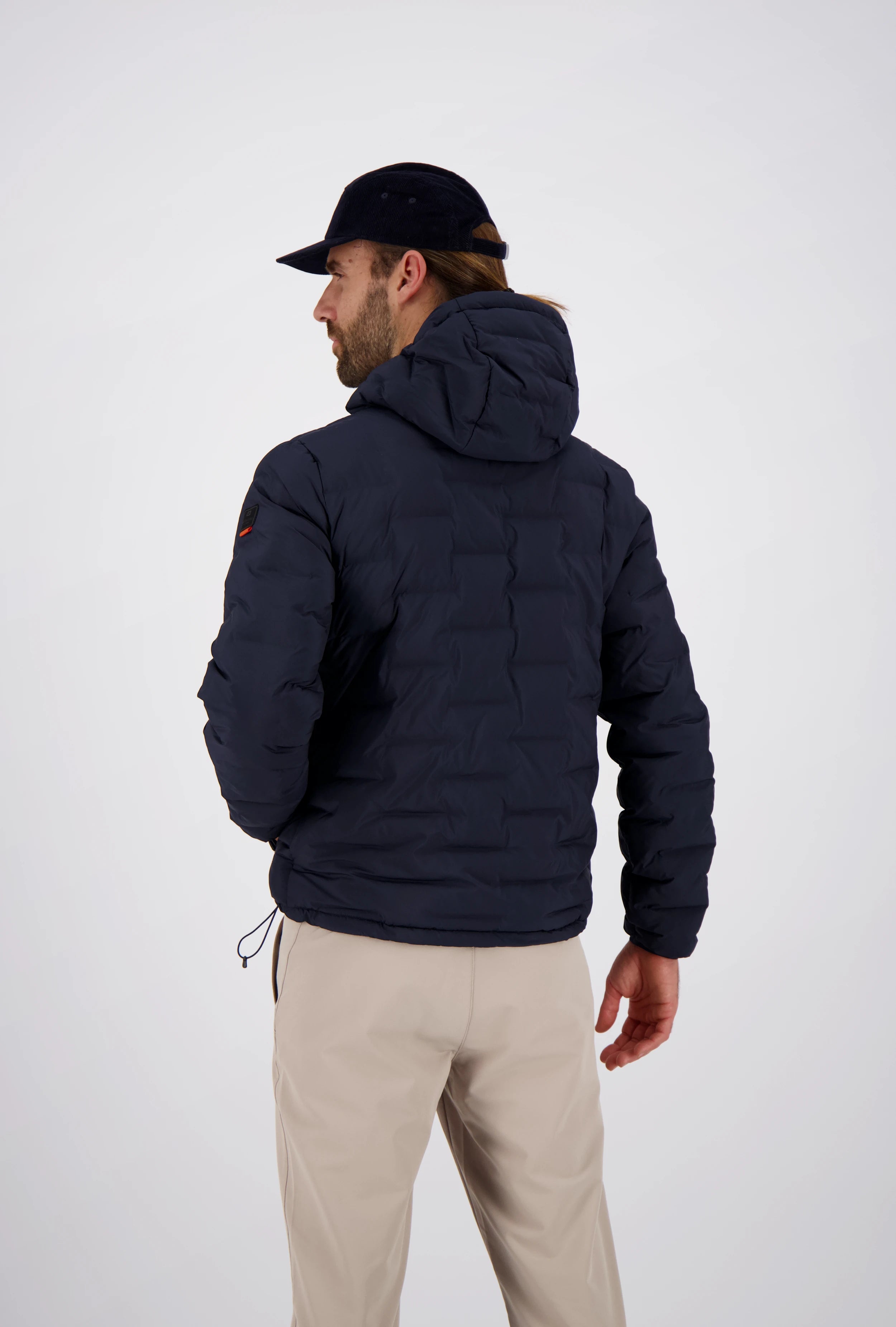 Rückenansicht eines Mannes mit dunkelblauer Cap in einer dunkelblauen Belowzero Herren Steppjacke mit Kapuze und Ärmel-Patch sowie beigefarbener Hose.
