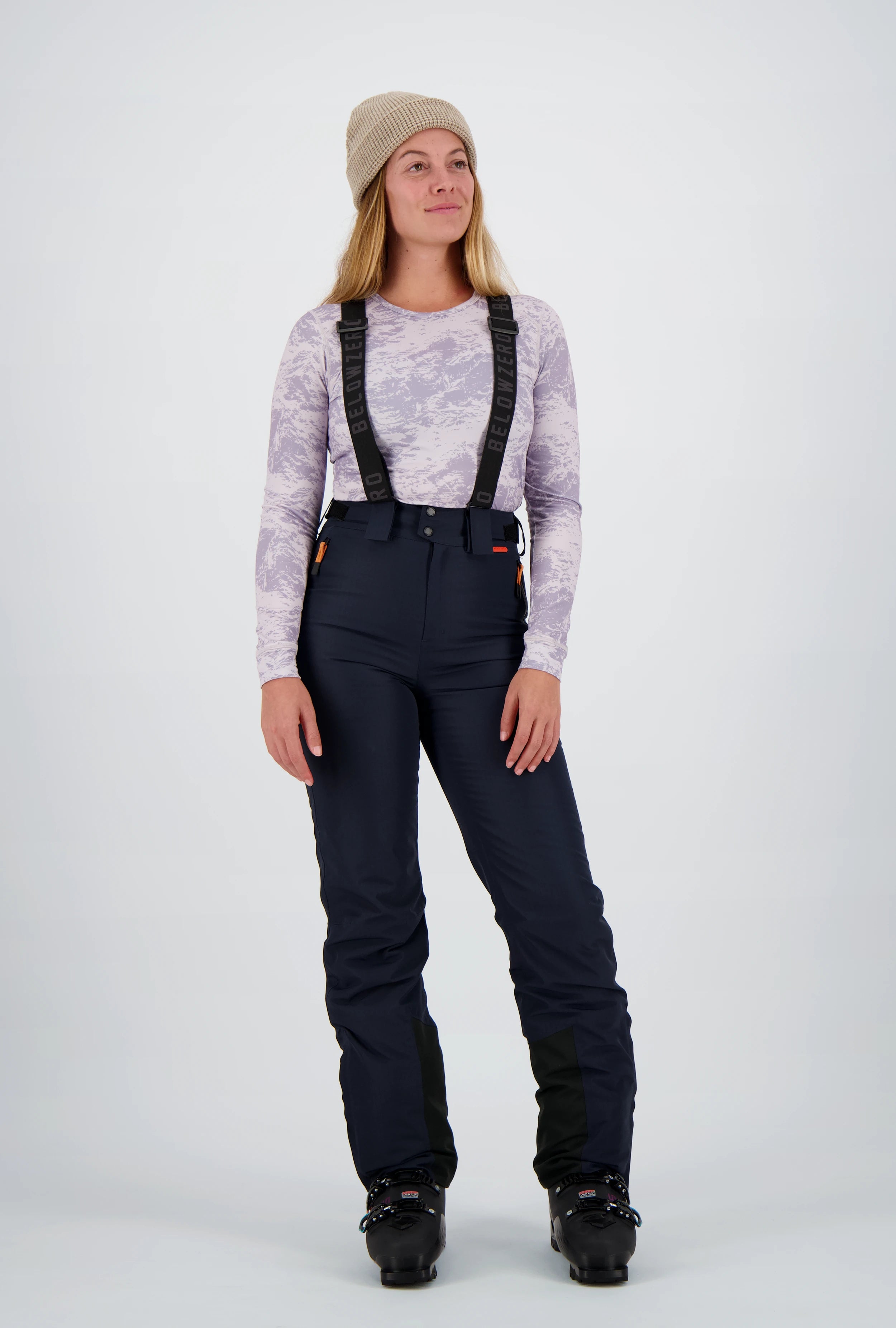Ganzkörperansicht einer Frau mit beigefarbener Beanie-Mütze in einer dunkelblauen Belowzero Damen Skihose mit verstellbaren Hosenträgern, gemustertem lila, violett Langarmshirt Longsleeve und schwarzen Skischuhen.