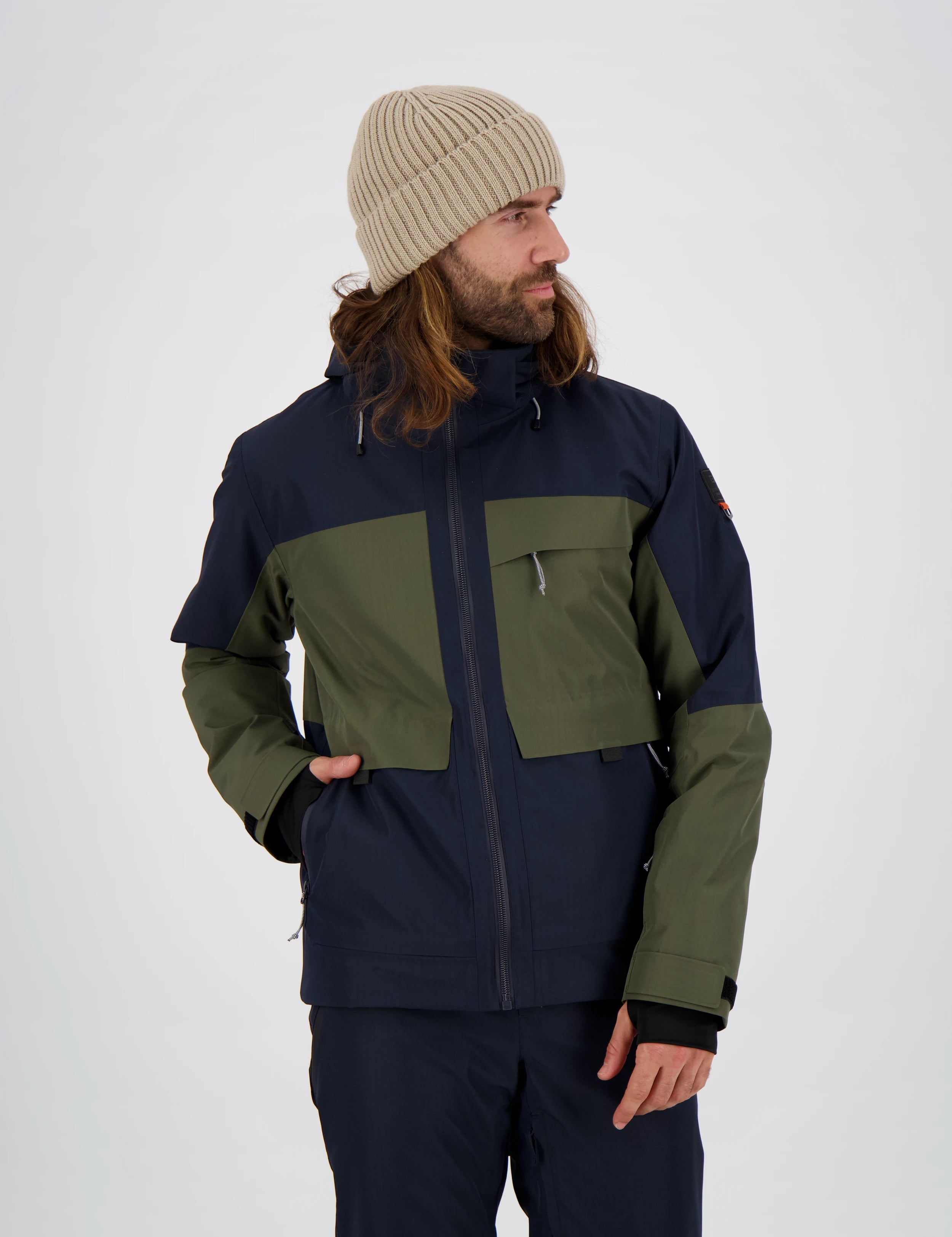 Mann trägt eine zweifarbige Belowzero Herren-Outdoorjacke in Dunkelblau und Olivgrün mit Kapuze und praktischen Taschen, kombiniert mit einer passenden dunkelblauen Outdoor-Hose und einer beigen Strickmütze.