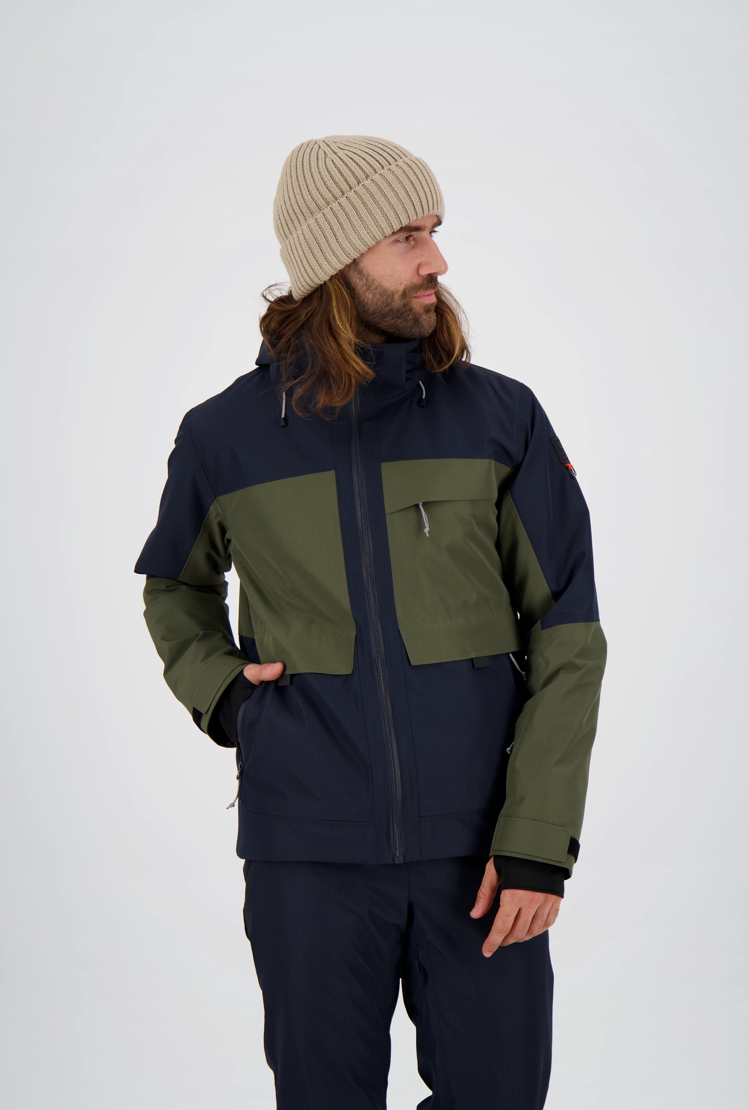 Mann trägt eine zweifarbige Belowzero Herren-Outdoorjacke in Dunkelblau und Olivgrün mit Kapuze und praktischen Taschen, kombiniert mit einer passenden dunkelblauen Outdoor-Hose und einer beigen Strickmütze.