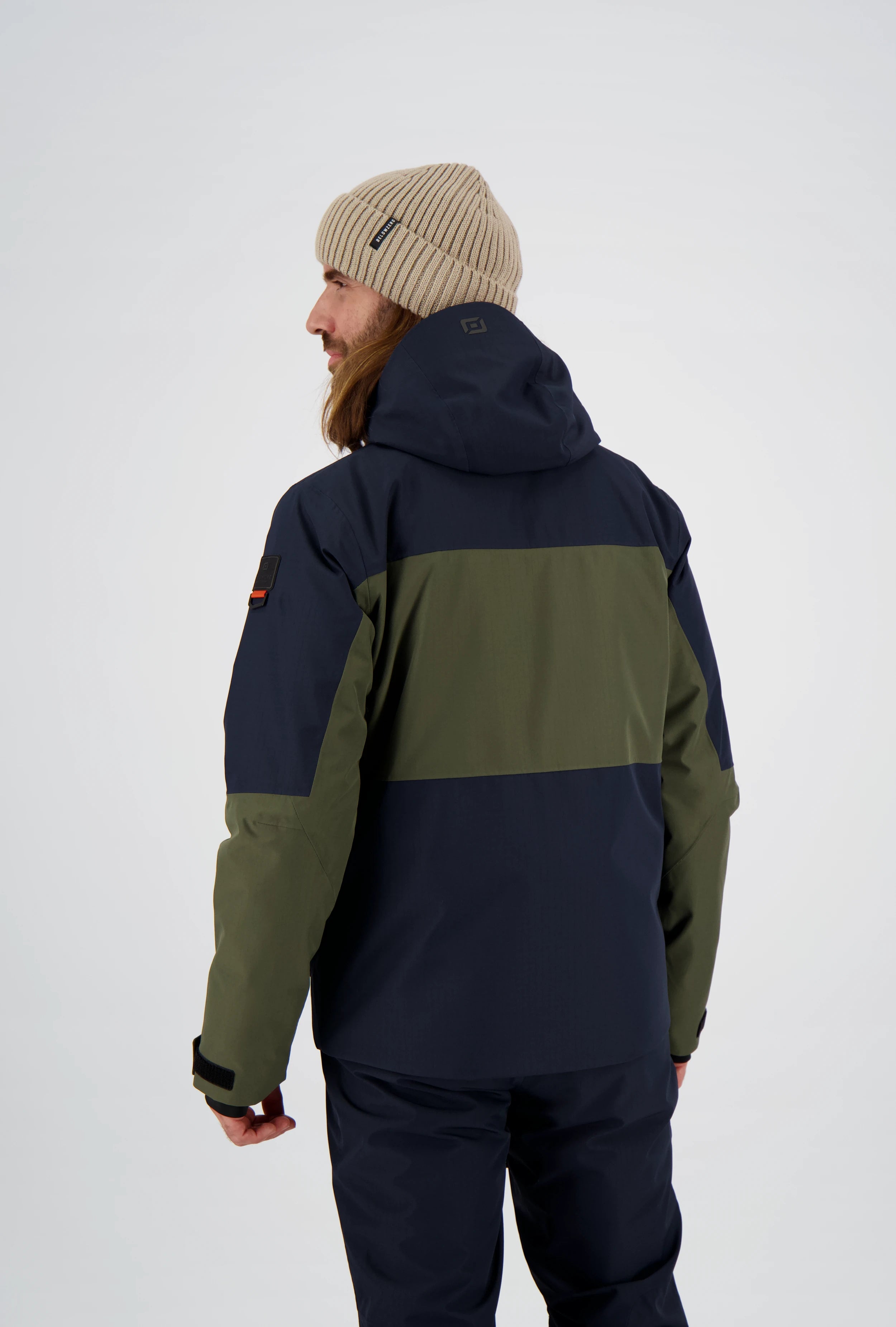 Rückansicht eines Mannes in einer zweifarbigen Belowzero Herren-Outdoorjacke in Dunkelblau und Olivgrün mit Kapuze, passender dunkler Hose und beiger Strickmütze.