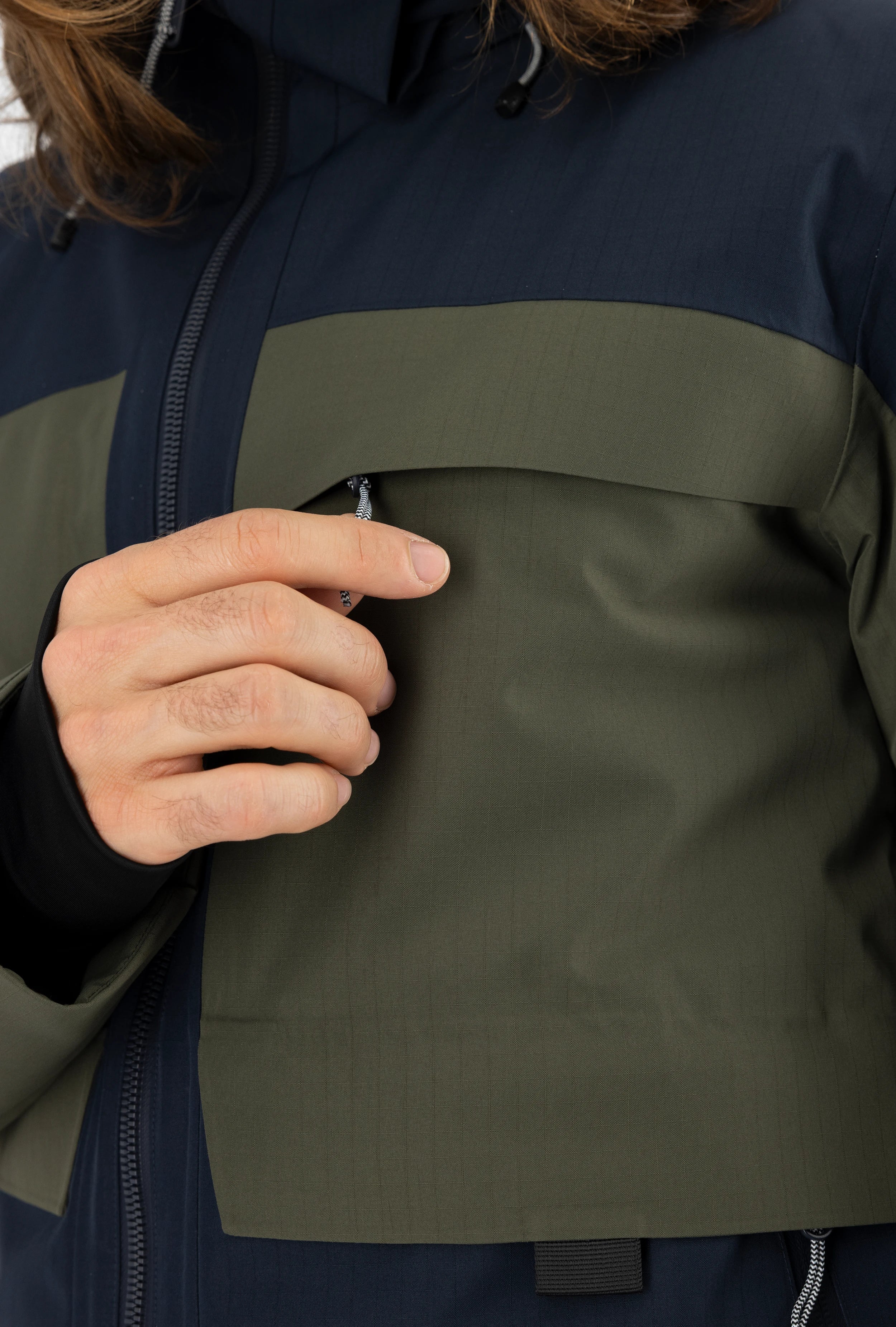 Detaillierte Nahaufnahme einer dunkelblau-grünen Belowzero Skijacke mit Verdeck für die Brusttasche, elastischen Pulswärmern am Ärmel und Kapuzenzug.