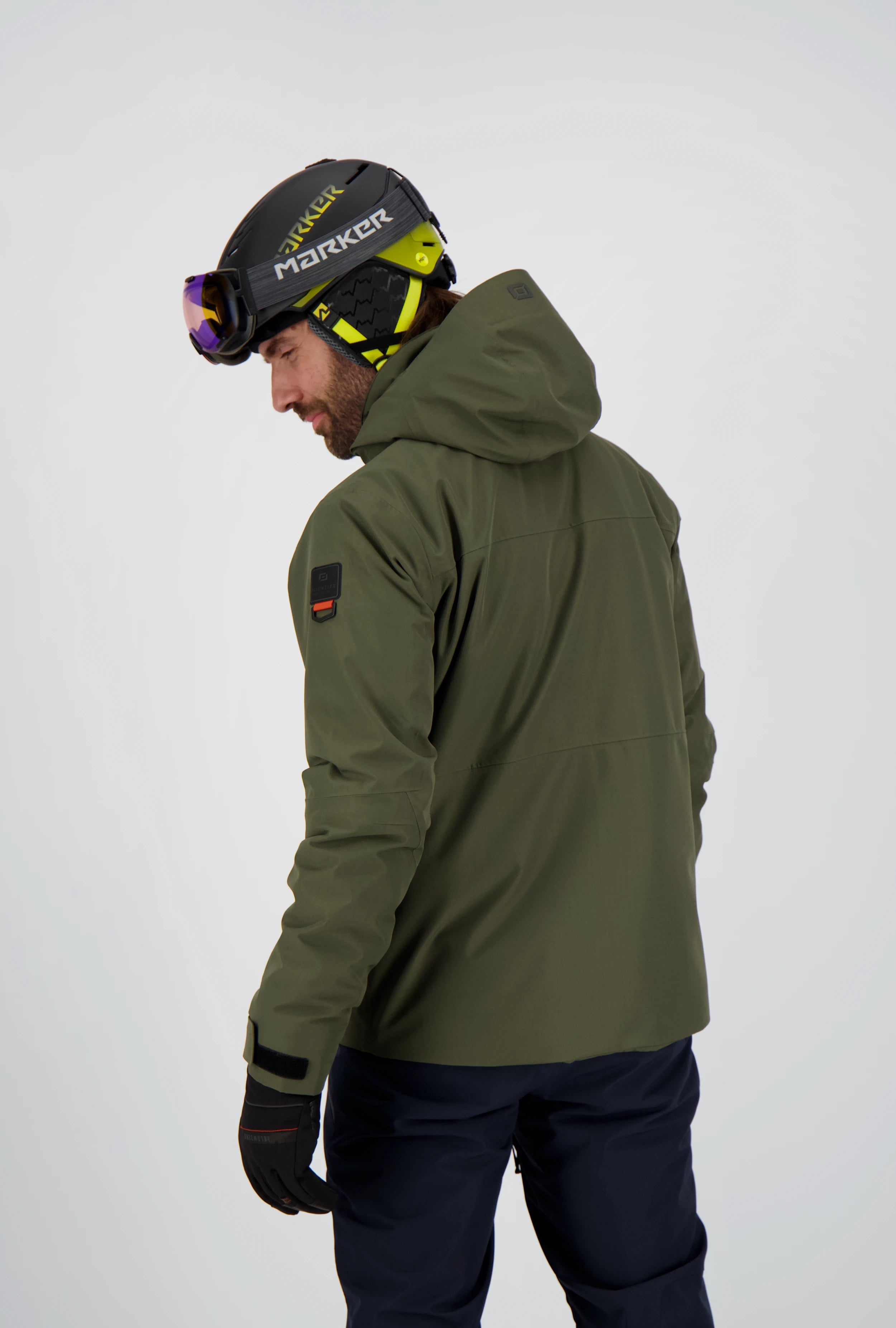 Rückansicht eines Mannes in einer olivgrünen Belowzero Skijacke mit Kapuze und Ärmel-Patch, dazu eine dunkle Skihose und Skihelm mit Brille.