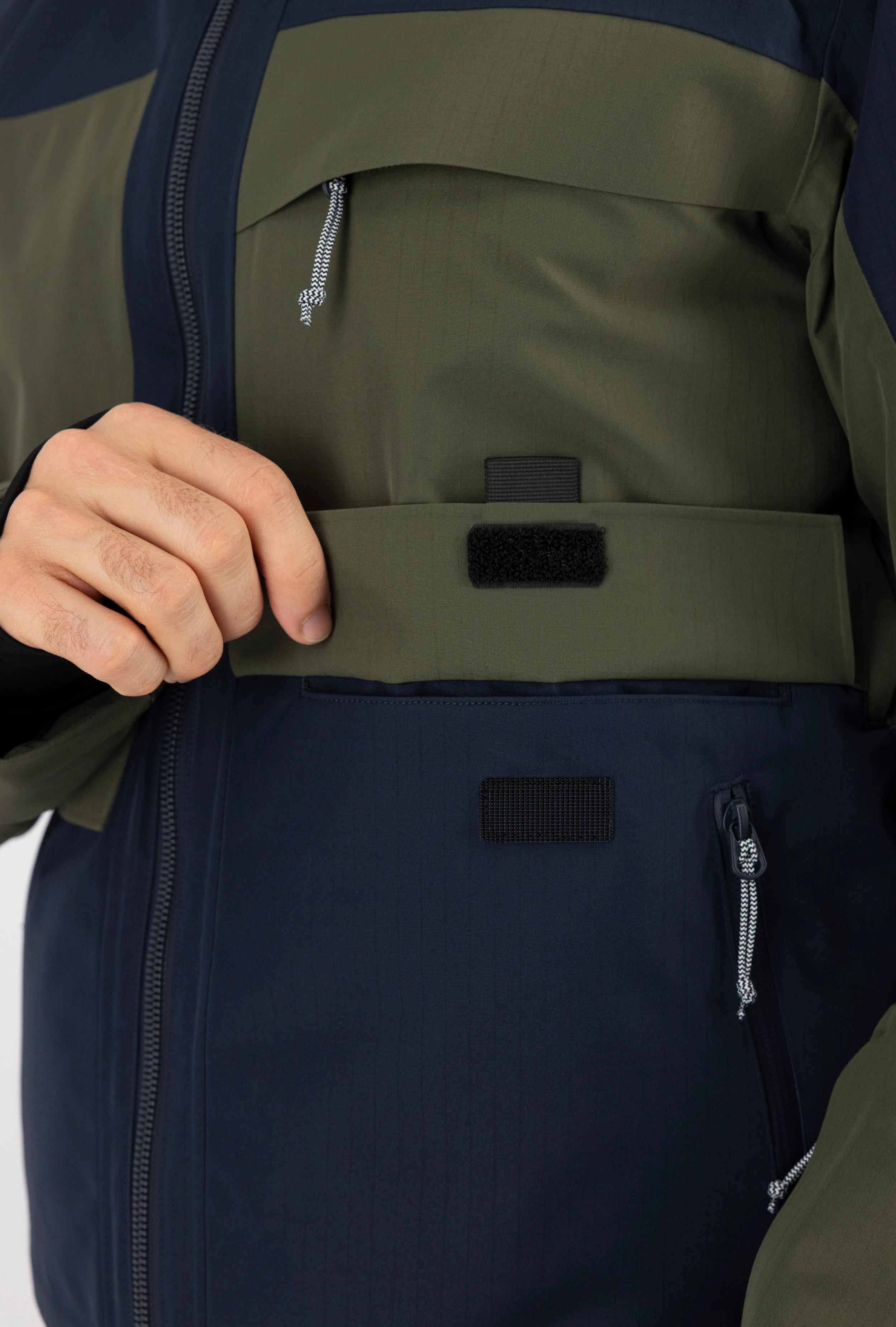 Detaillierte Nahaufnahme einer zweifarbigen Belowzero Skijacke in Dunkelblau und Olive mit Fokus auf die Windschutzleiste mit Klettverschluss und Taschen-Details.