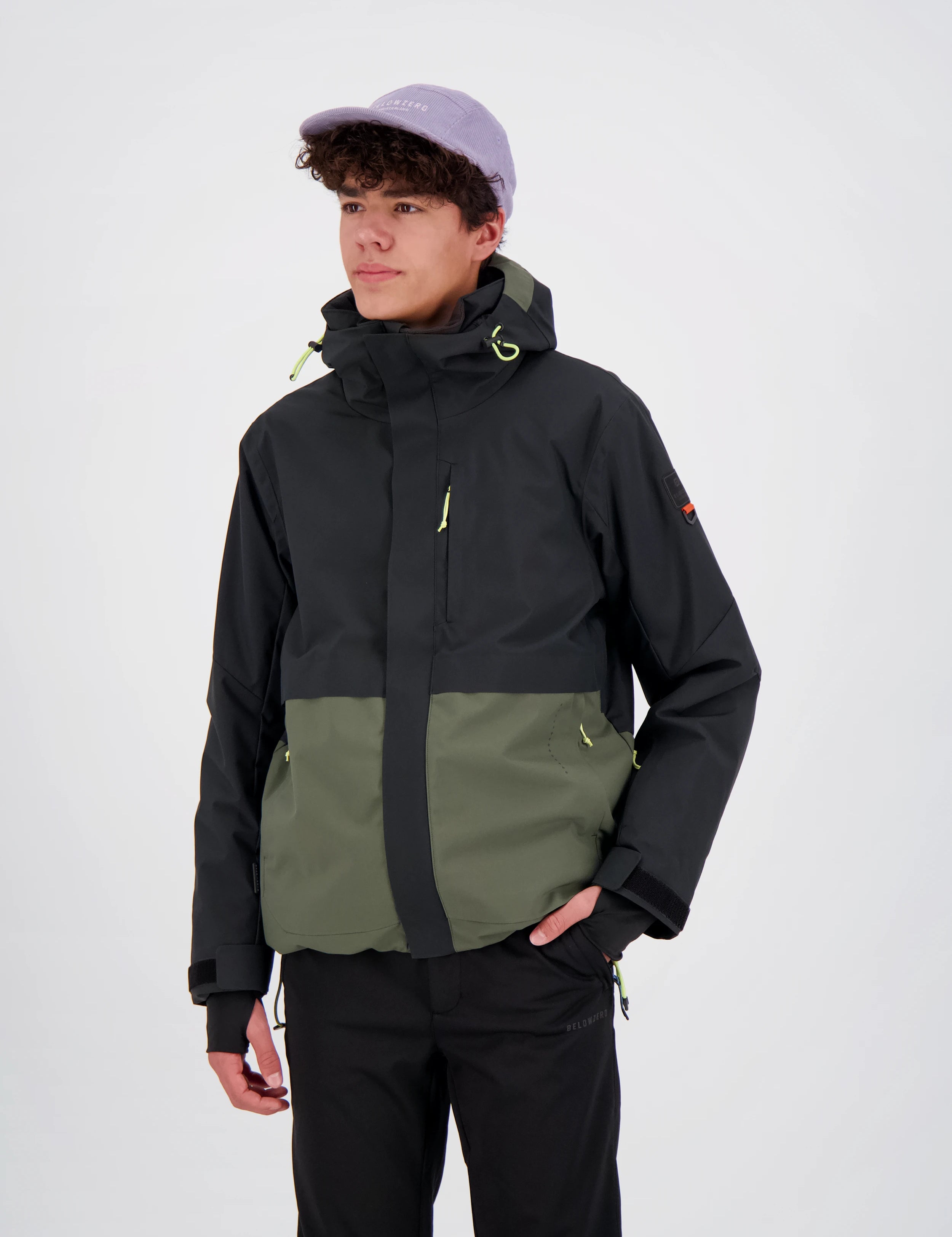 Junger Mann trägt eine zweifarbige Belowzero Outdoor-Skijacke in Schwarz und Olivgrün mit Kapuze, integrierten Handgamaschen und verstellbaren Bündchen, kombiniert mit einer schwarzen Funktionshose und einer hellvioletten Cap.