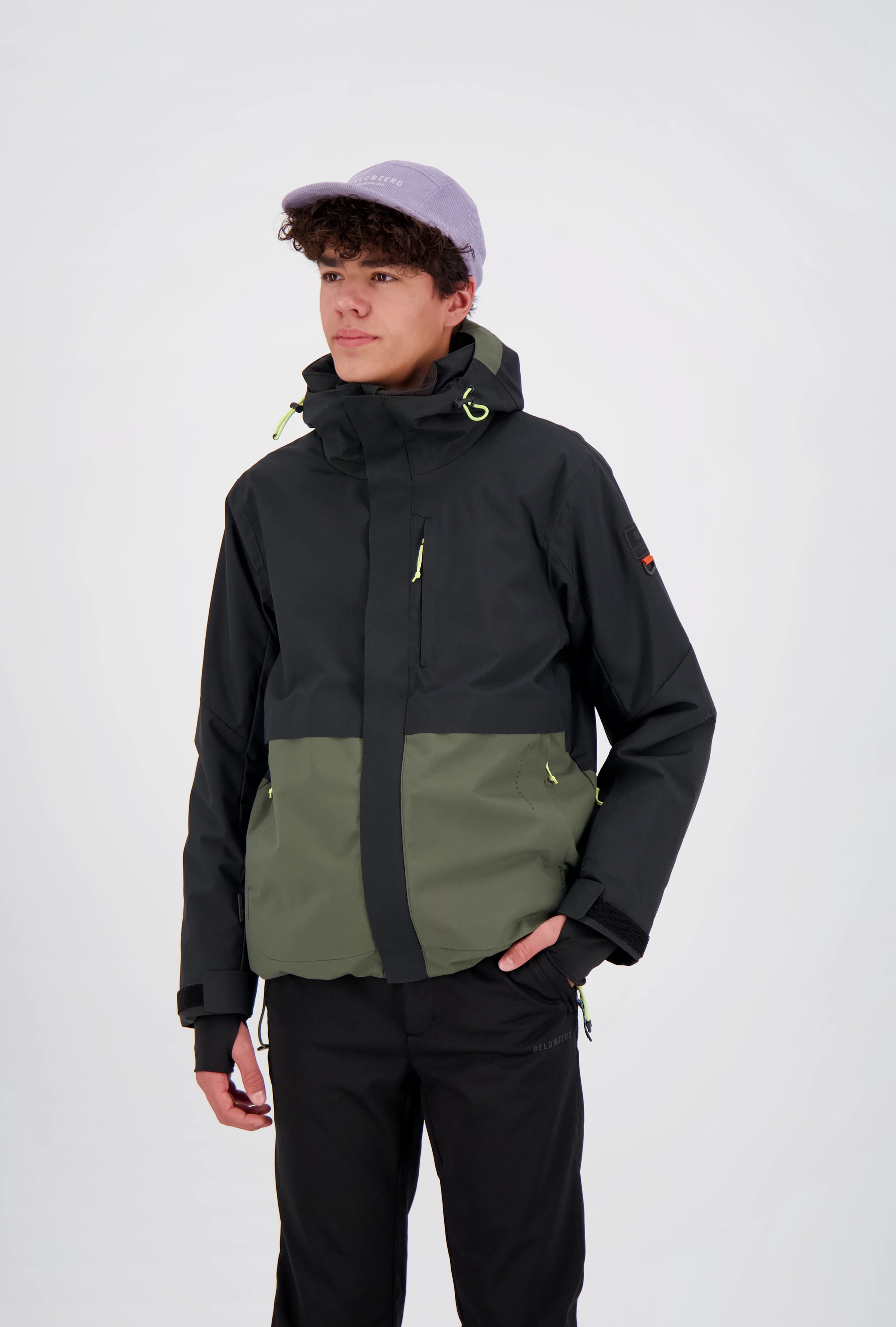 Junger Mann trägt eine zweifarbige Belowzero Outdoor-Skijacke in Schwarz und Olivgrün mit Kapuze, integrierten Handgamaschen und verstellbaren Bündchen, kombiniert mit einer schwarzen Funktionshose und einer hellvioletten Cap.