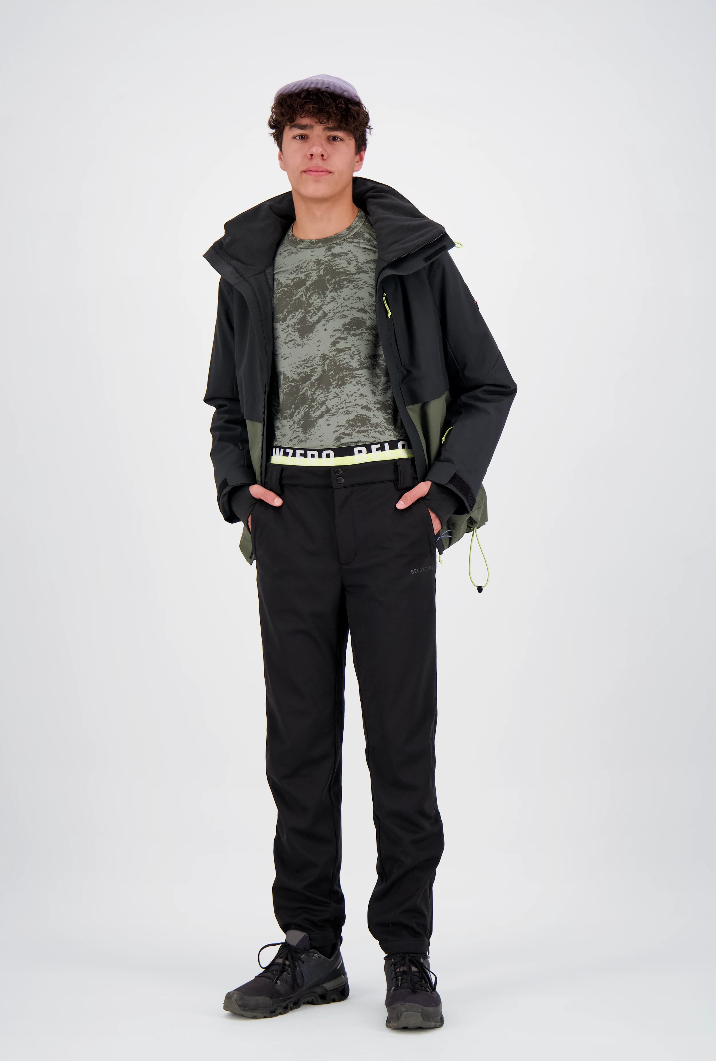 Junger Mann zeigt ein dreiteiliges Belowzero Herren-Outdoor-Outfit: Eine geöffnete zweifarbige Jacke in Schwarz und Olivgrün, ein grün gemustertes Funktionsshirt (Baselayer) mit sichtbarem Belowzero-Logo-Bund und eine schwarze Funktionshose.