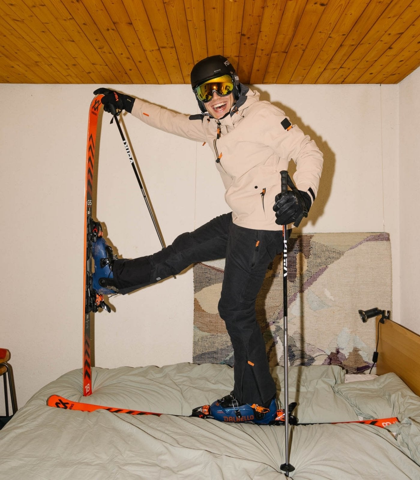Ein Herr trägt Belowzero Bekleidung und die gesamte Skiausrüstung. Dabei steht er auf einem Bett in einem Zimmer.