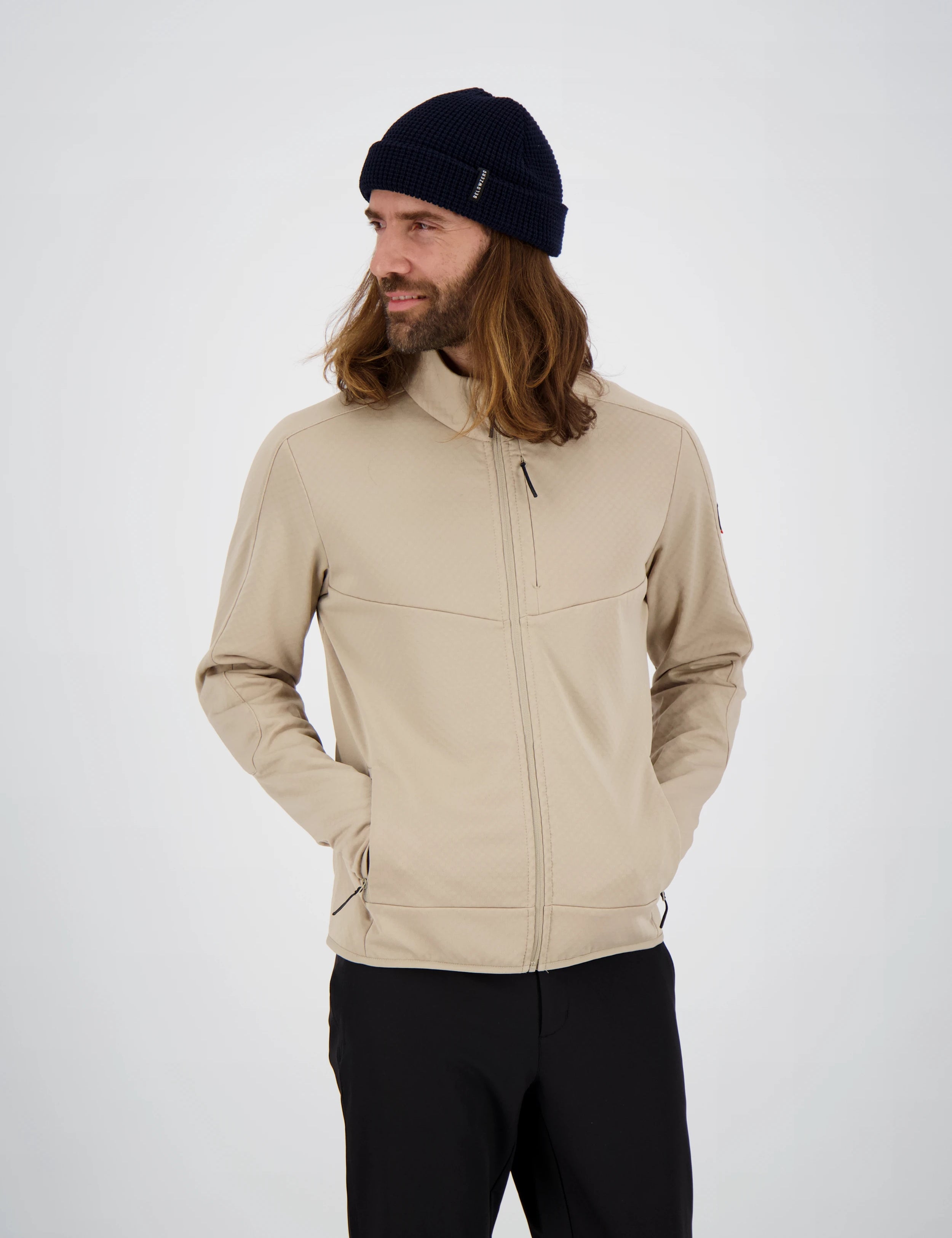 Frontansicht eines Mannes mit langen Haaren und dunkelblauer Beanie-Mütze in einer beigefarbenen Belowzero Herren Fleecejacke Midlayer mit Brusttasche und schwarzer Hose.