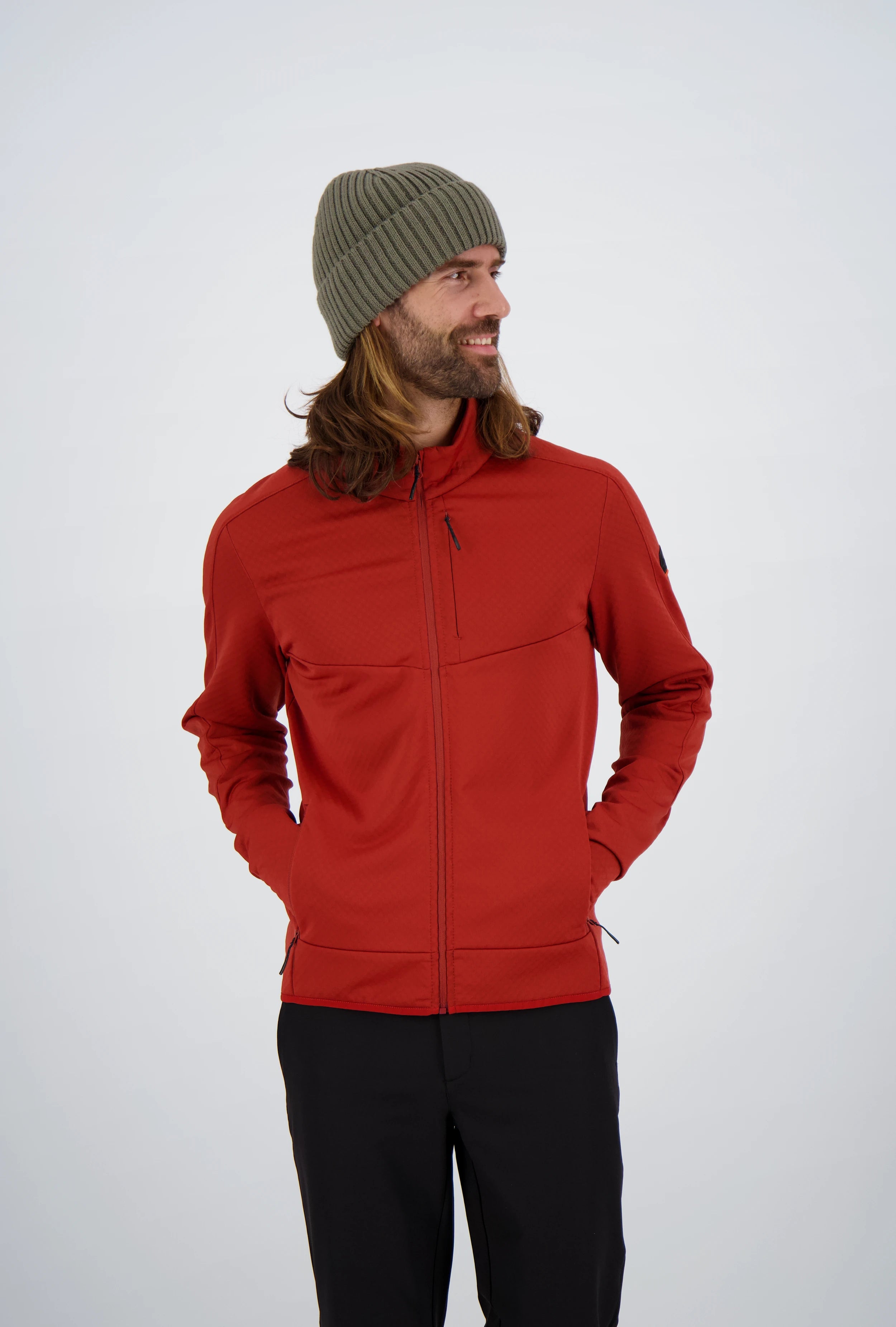 Frontansicht eines Mannes mit langen Haaren und olivgrüner Beanie-Mütze in einer roten Belowzero Herren Fleecejacke Midlayer mit Brusttasche und schwarzer Hose