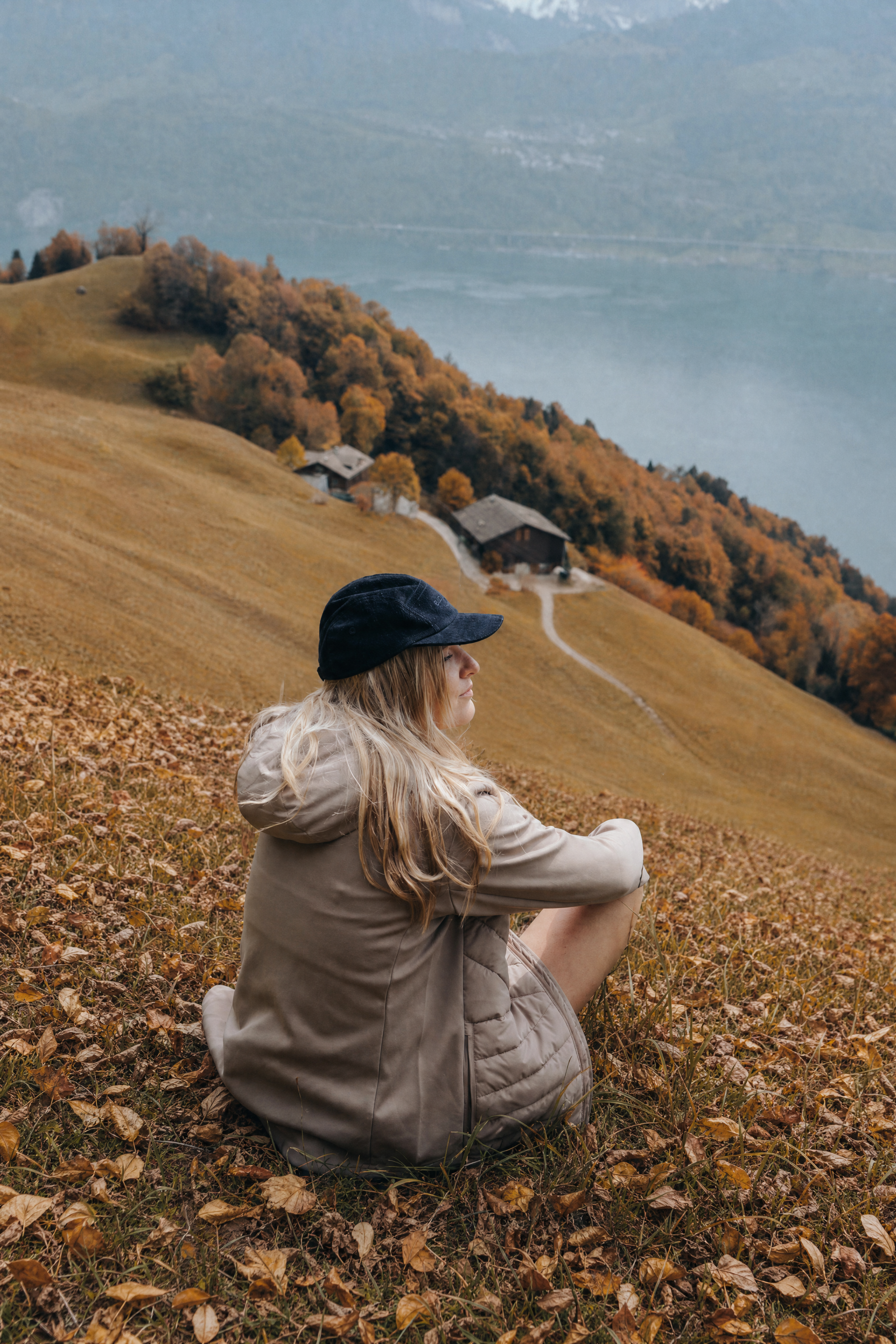 Frau sitzt von hinten in einer beigen Belowzero Outdoor-Kapuzenjacke mit gesteppten Details und dunkler Mütze auf einer Wiese mit Blick auf Berglandschaft und See.