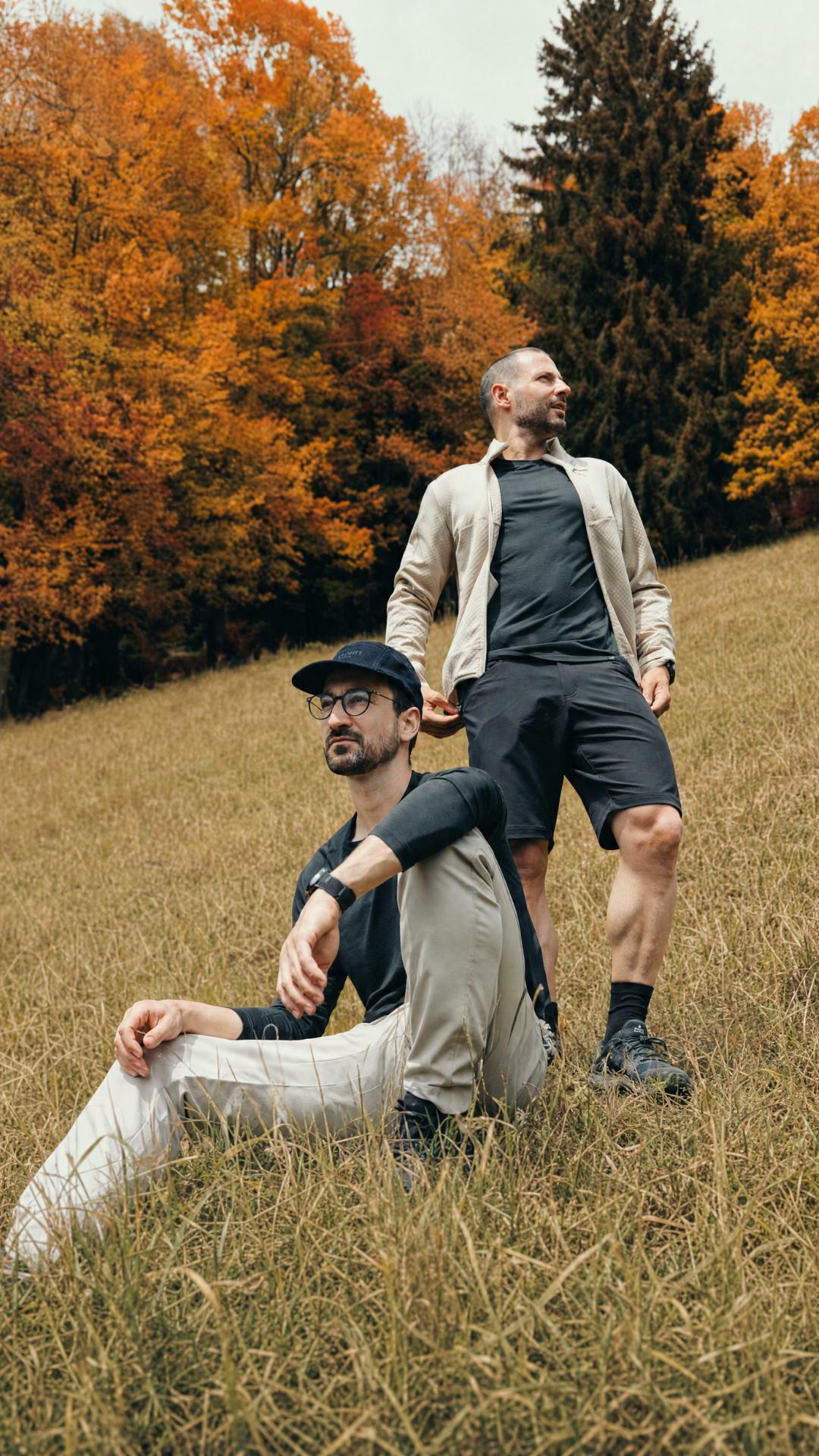 Zwei Männer in funktioneller Belowzero Outdoor-Bekleidung für Herren in der Herbstlandschaft: Stehender Mann trägt eine beige Fleecejacke, dunkles Funktionsshirt und schwarze Shorts; sitzender Mann trägt ein dunkles Langarm-Funktionsshirt, eine helle Outdoor-Hose und eine schwarze Mütze.