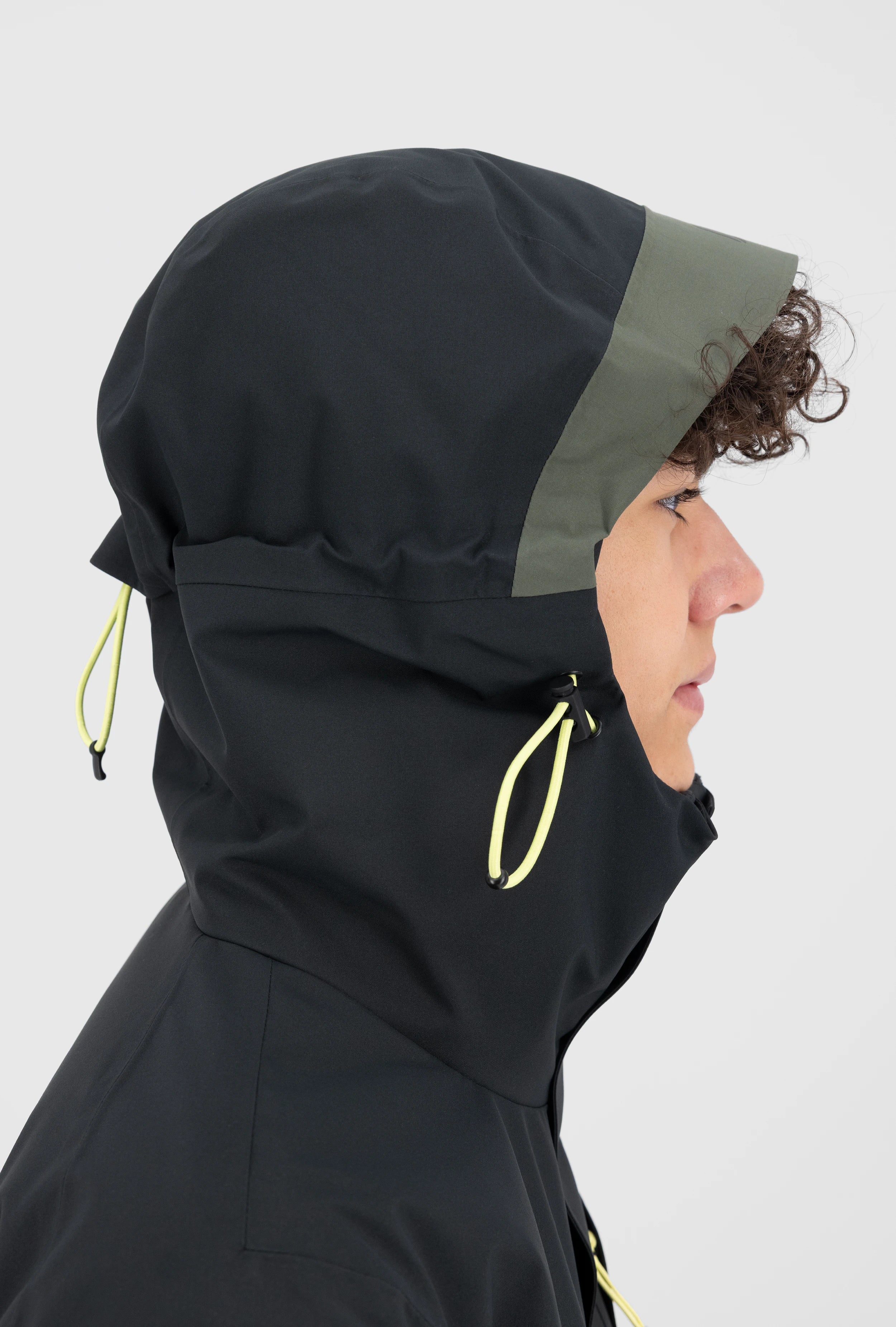 Detaillierte Seitenansicht einer schwarzen Belowzero Herren Outdoorjacke mit verstellbarer Kapuze, grünem Schild, hellgelben Kordelzügen und hochgeschlossenem Kragen für optimalen Wetterschutz.