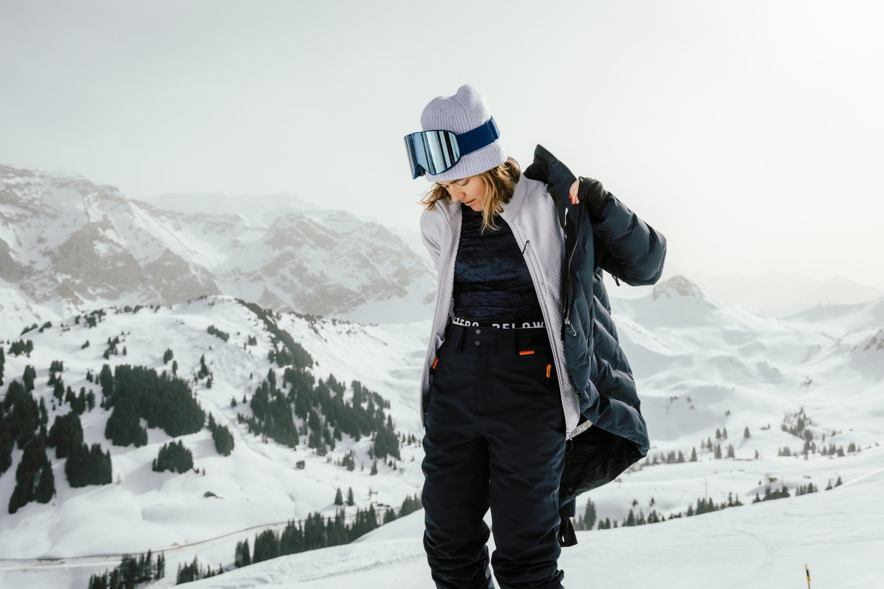 Frau in Belowzero Skibekleidung mit Skibrille und Mütze vor verschneitem Bergpanorama
