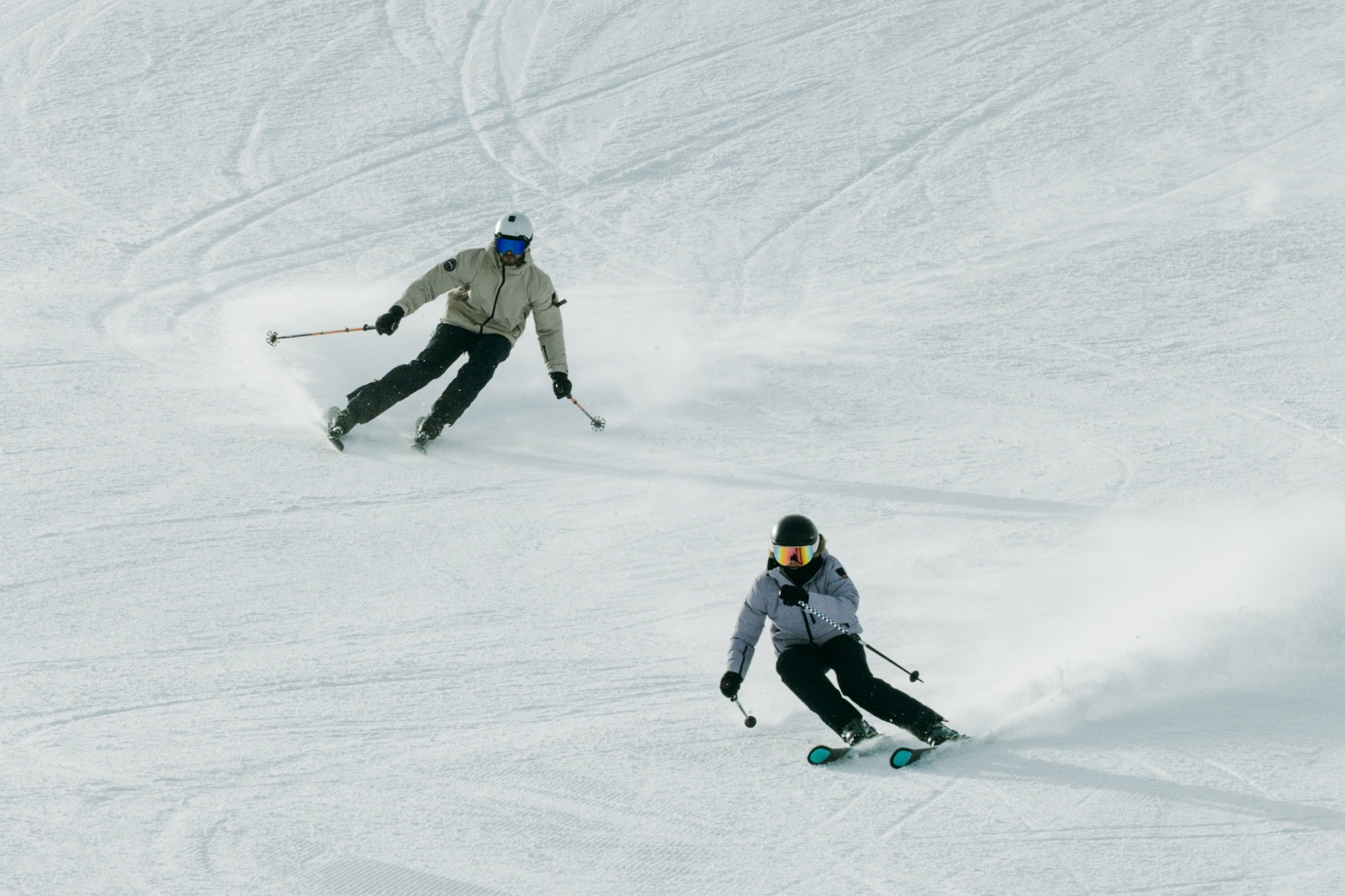 Zwei Personen in Belowzero Skibekleidung carven eine Piste hinunter, Schnee stiebt auf